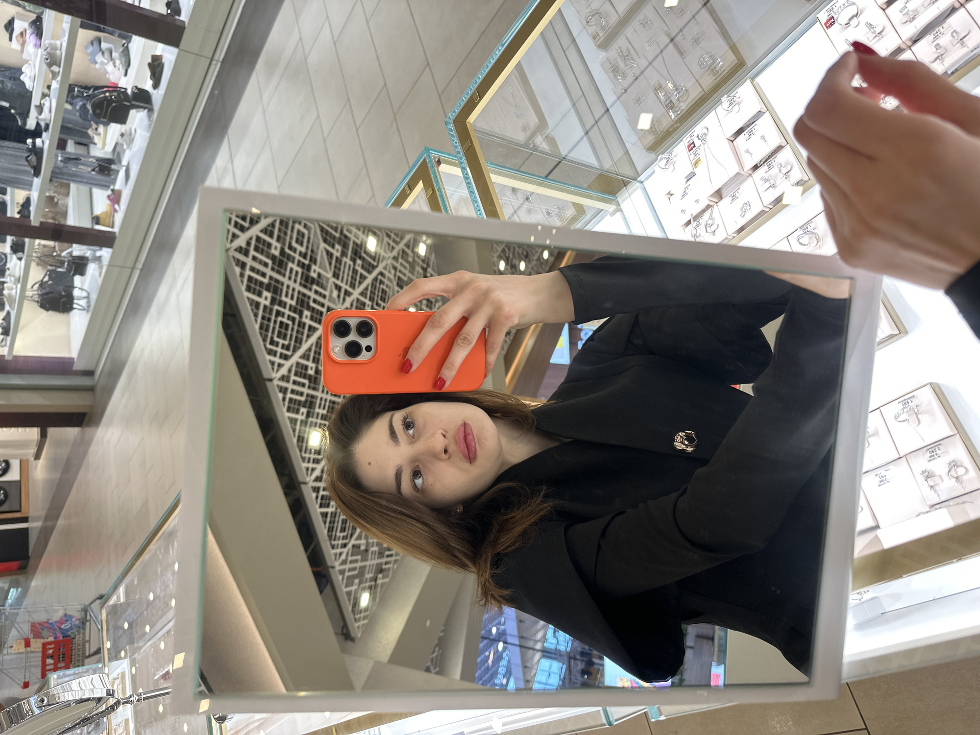 The image depicts a mirror selfie taken by a woman inside a jewelry store, situated within a shopping mall in Yekaterinburg, Russia. The subject is a woman with brown hair, wearing a black blazer-style garment with a gold-toned decorative button. Her left hand, visible in the mirror, holds an orange-cased smartphone with a multi-lens camera array, positioned to capture her reflection. Her fingernails are painted red. Her right hand is partially visible in the foreground, outside the mirror's frame, also with red nail polish, positioned over a jewelry display case.
The environment consists of several glass display cases with gold and light blue trim, illuminated internally, showcasing numerous silver-colored jewelry items such as rings, pendants, and earrings, each tagged with small white price labels indicating Russian Rubles (e.g., "1,990 ₽"). The floor is composed of light-colored, square tiles. Reflections in the background reveal a spacious shopping mall environment, including other retail storefronts displaying shoes and handbags, and a ceiling with a geometric grid pattern. A red-accented shopping cart is partially visible in a distant reflection.