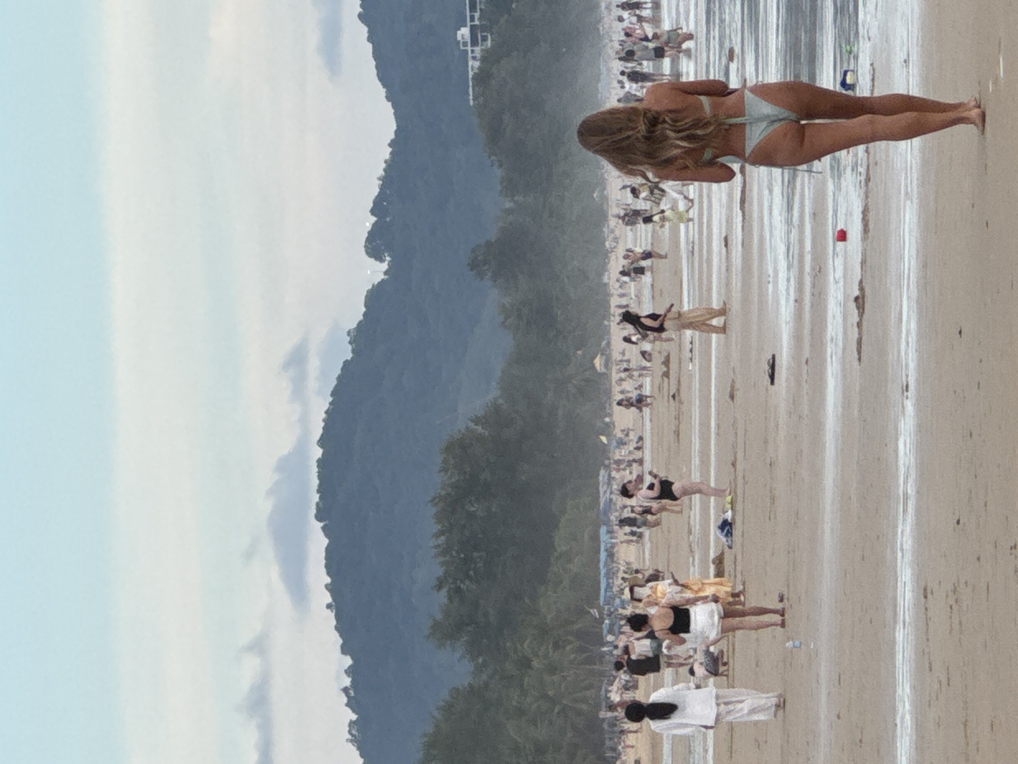 This is a full-frame, eye-level, medium shot of Karon Beach in Karon, Thailand, on a hazy day. The sand is light brown, wet in some areas, and littered with debris. The water is a light blue-grey, with small waves lapping at the shore. In the foreground, a woman with long, wavy blonde hair walks away from the camera, wearing a light blue bikini. Behind her, the beach is crowded with people. Several individuals are walking, standing, or sitting on the sand. Among them, a woman in a white outfit with her back to the camera stands near the left edge. Further back, a woman in a black bikini is visible. The background features a dense line of green trees, followed by a large, dark green, mist-covered mountain range under a pale blue sky. A building is visible on the mountainside.