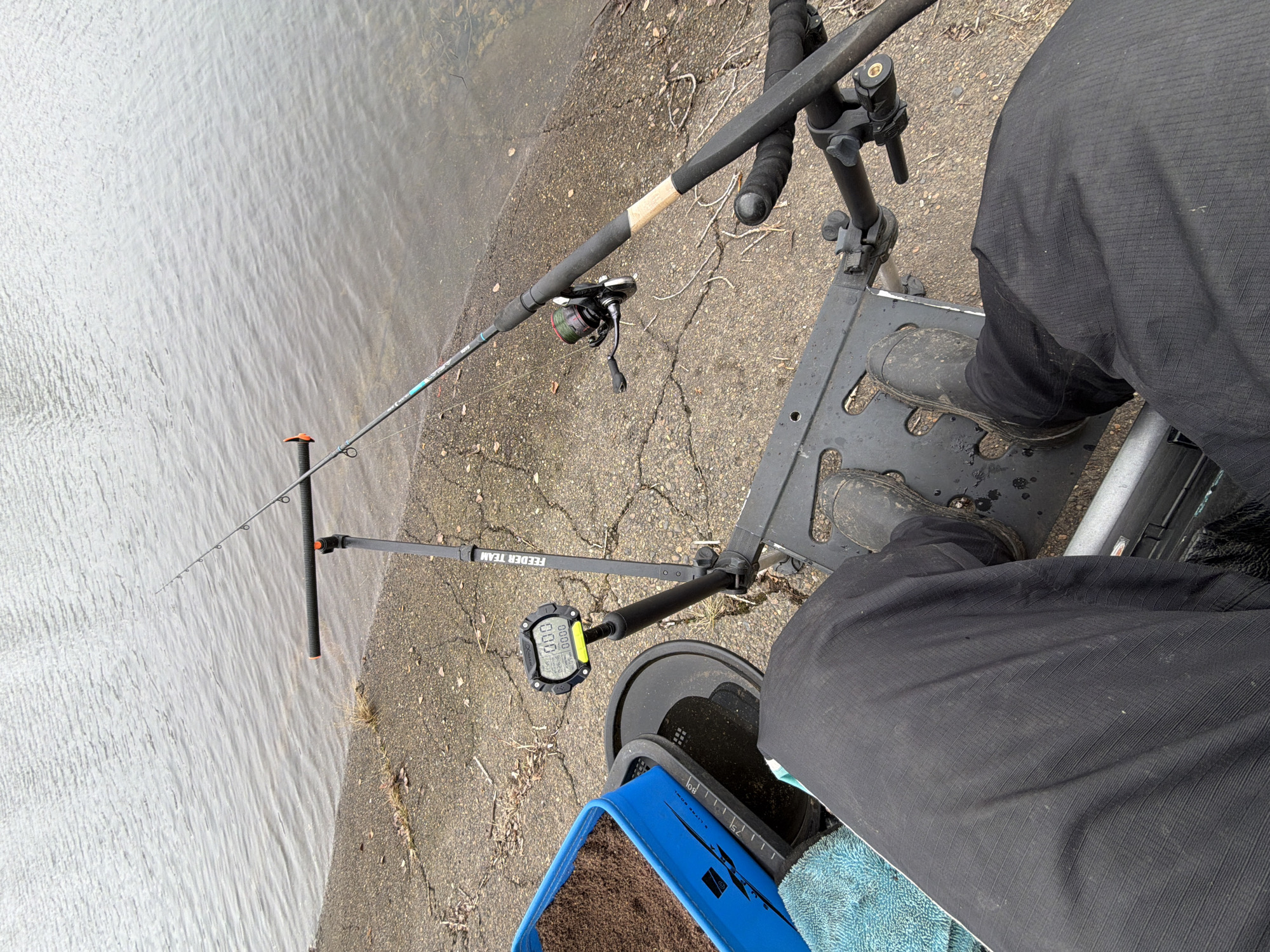 A high-angle, point-of-view photograph shows a person fishing in an urban environment. The person is seated, wearing dark pants and muddy boots on a platform. Two fishing rods are visible: one with a reel and another resting on a rod support. A Feeder Team electronic device is nearby, likely for measuring distance. A blue container with a towel is present. The background shows water and a concrete shoreline. The specific location in the city of Chaikovsky, Russia cannot be determined without further information.