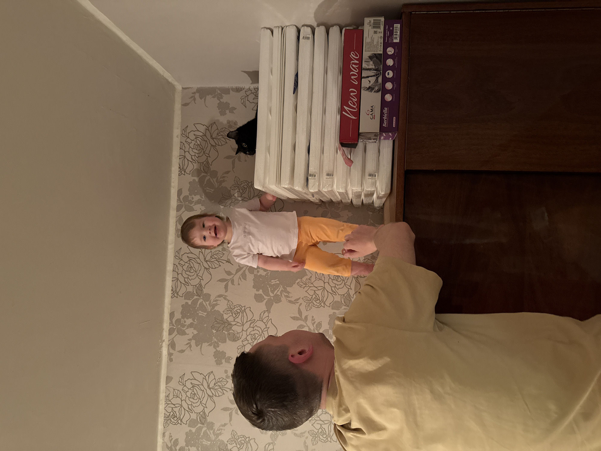 The image shows a man, a child, and a black cat in an indoor setting. The man, wearing a light yellow shirt, is facing the camera. The child, wearing a white shirt and yellow pants, is lying on the floor. A black cat peeks out from behind a stack of white objects. A wooden cabinet is visible on the right side of the image. The floor has a floral wallpaper pattern. The location is possibly a room in a building in Krapivka, Russia.