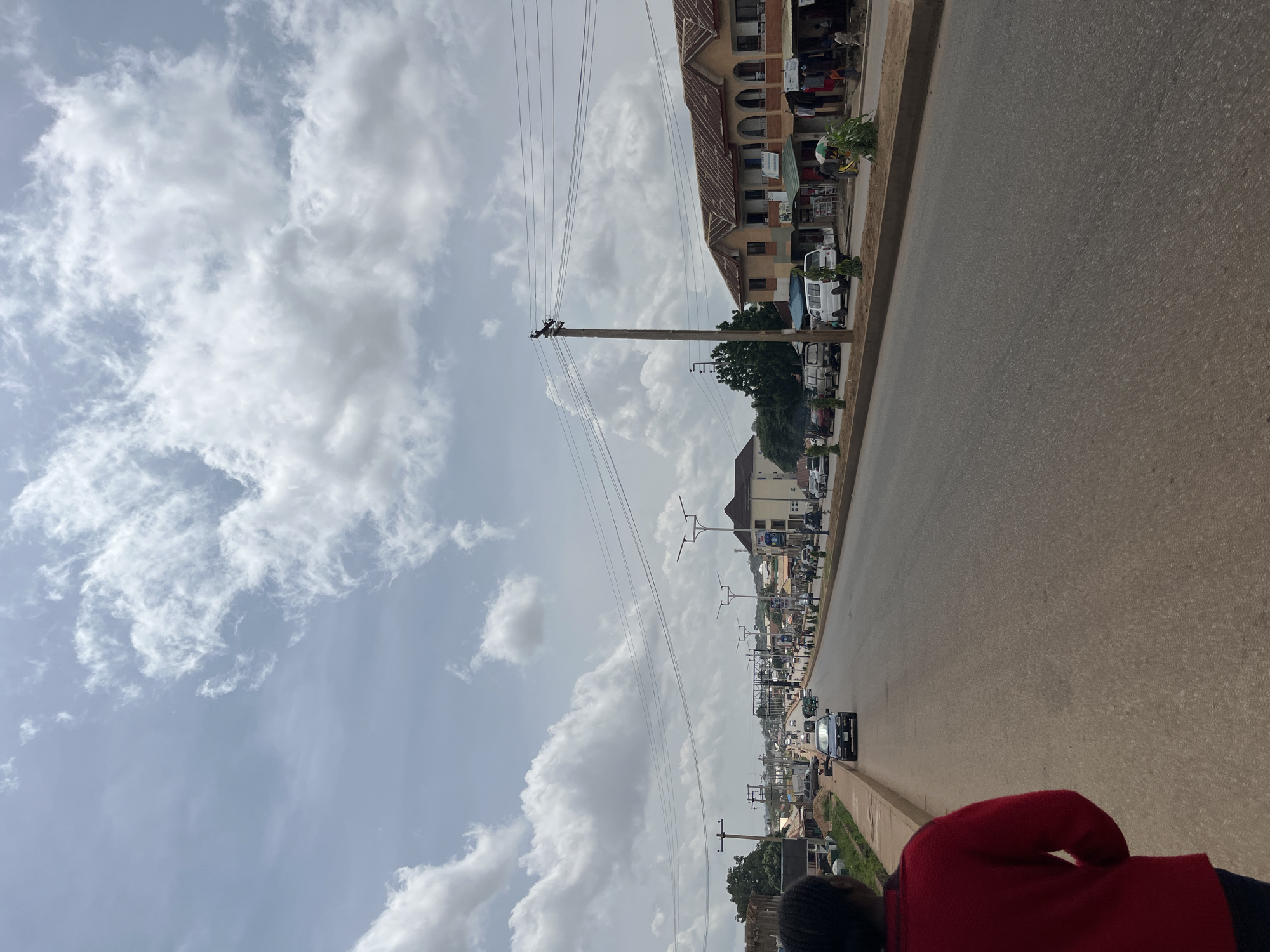 The image depicts a street scene in Bukuru, Nigeria, likely on a partly cloudy day. A person in a red shirt is walking on the side of a road. The road appears to be a main thoroughfare with multiple lanes and moderate traffic, including cars and vans. Along the right side of the road, there are buildings that appear to be shops and residential structures, some with awnings and merchandise displayed outside. Power lines and streetlights are visible along the road. In the distance, the road extends into a more built-up area with more buildings and infrastructure. The sky is filled with scattered white clouds.