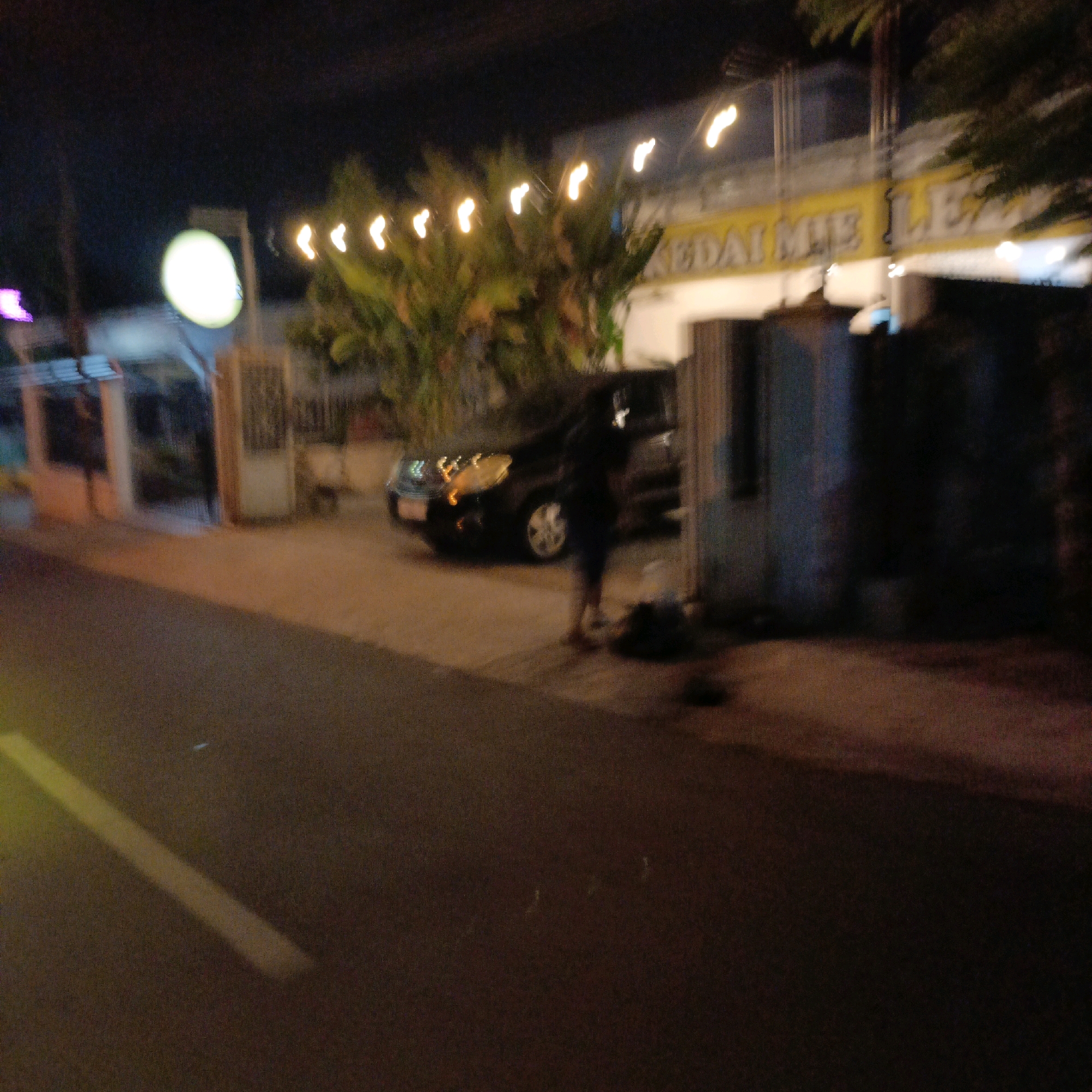A dark, out-of-focus nighttime image captured in Salatiga, Indonesia, shows a street scene in front of a building with a yellow sign. The sign has text on it that appears to read "KEDAI MIE LE..." which likely refers to a noodle shop. String lights are visible along the top of the building. A dark-colored car is parked on the right side of the street, and a person in dark clothing is standing near it. The street in the foreground has a faint yellow line marking, indicating it is a road. The overall atmosphere is dimly lit, suggesting it is late at night.