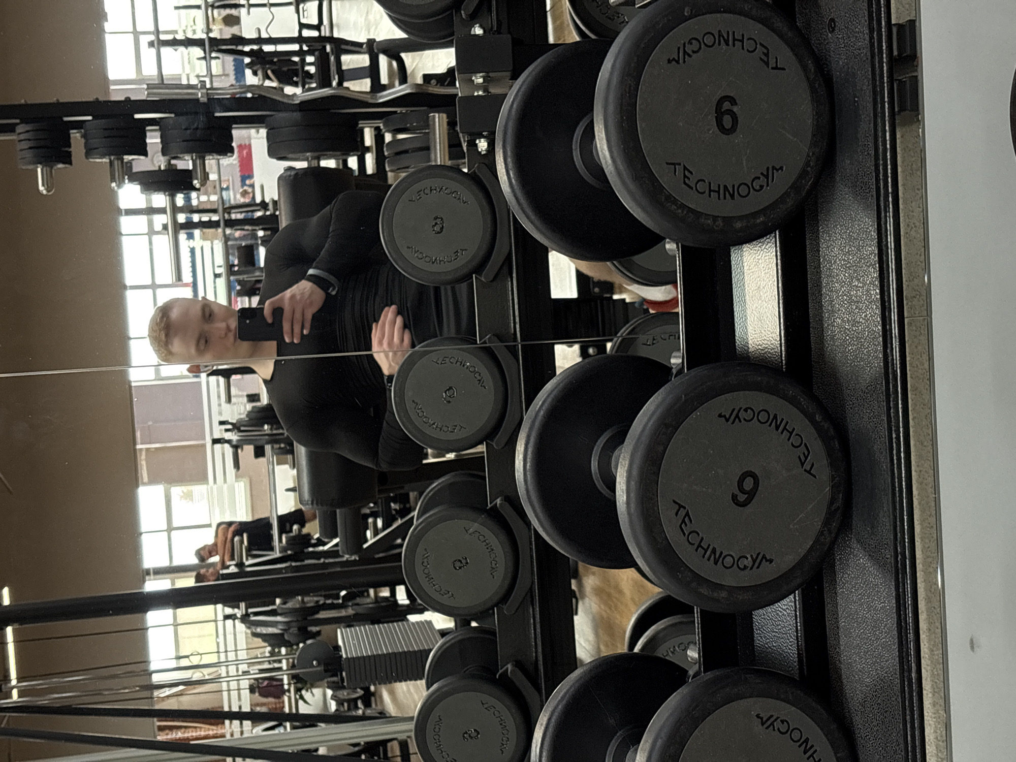 This image, captured in a gym in Chaikovsky, Russia, depicts a reflection of a young man taking a mirror selfie. The man, with short blonde hair and wearing a black long-sleeved top, is seen holding a black smartphone to photograph his reflection. He stands in front of a robust black rack holding various dumbbells. Several dumbbells are prominently displayed, featuring the brand name "TECHNOGYM" and specific weight numbers, including "6", "8", and "9". In the background of the reflection, large windows allow natural light to illuminate the space, suggesting it is daytime. The reflection also reveals other gym equipment, such as weight plates on barbells and machine frames, along with a faint outline of another person, indicating an active fitness environment. The scene effectively captures a personal moment within a contemporary gym setting.