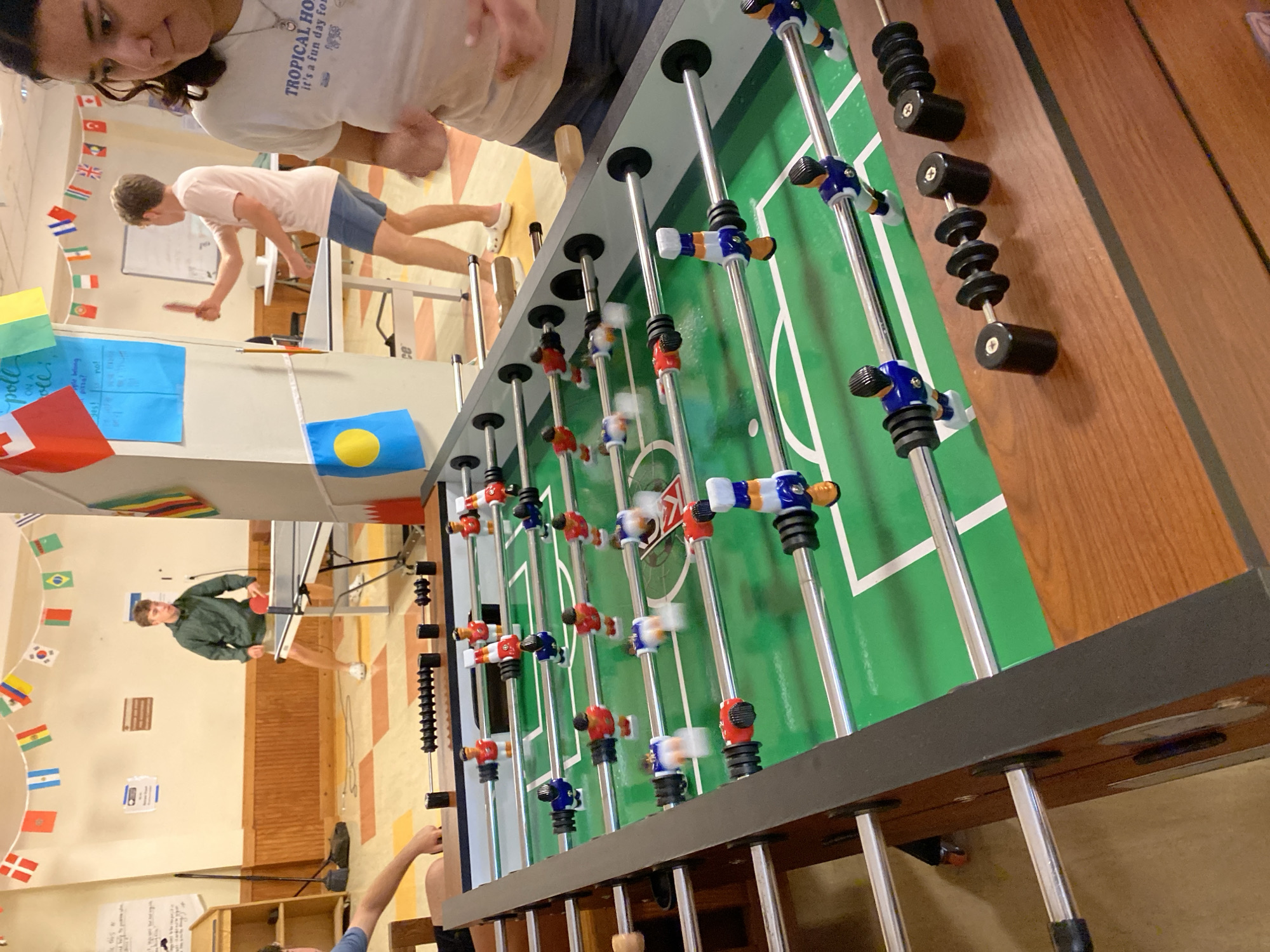 In Washington Township, USA, a foosball table dominates the foreground of an indoor recreation area. The table features a green playing surface with white markings and rows of miniature red, blue, and white player figures on metal rods. In the background, two young people are engaged in a table tennis match. One, wearing a green hooded jacket and dark shorts, stands to the left of a table tennis table, holding a red paddle. The other, in a pink t-shirt and blue shorts, is on the right, also with a paddle. The room is decorated with a string of international flags and various posters on the walls. The floor has a checkerboard pattern of orange and yellow tiles.