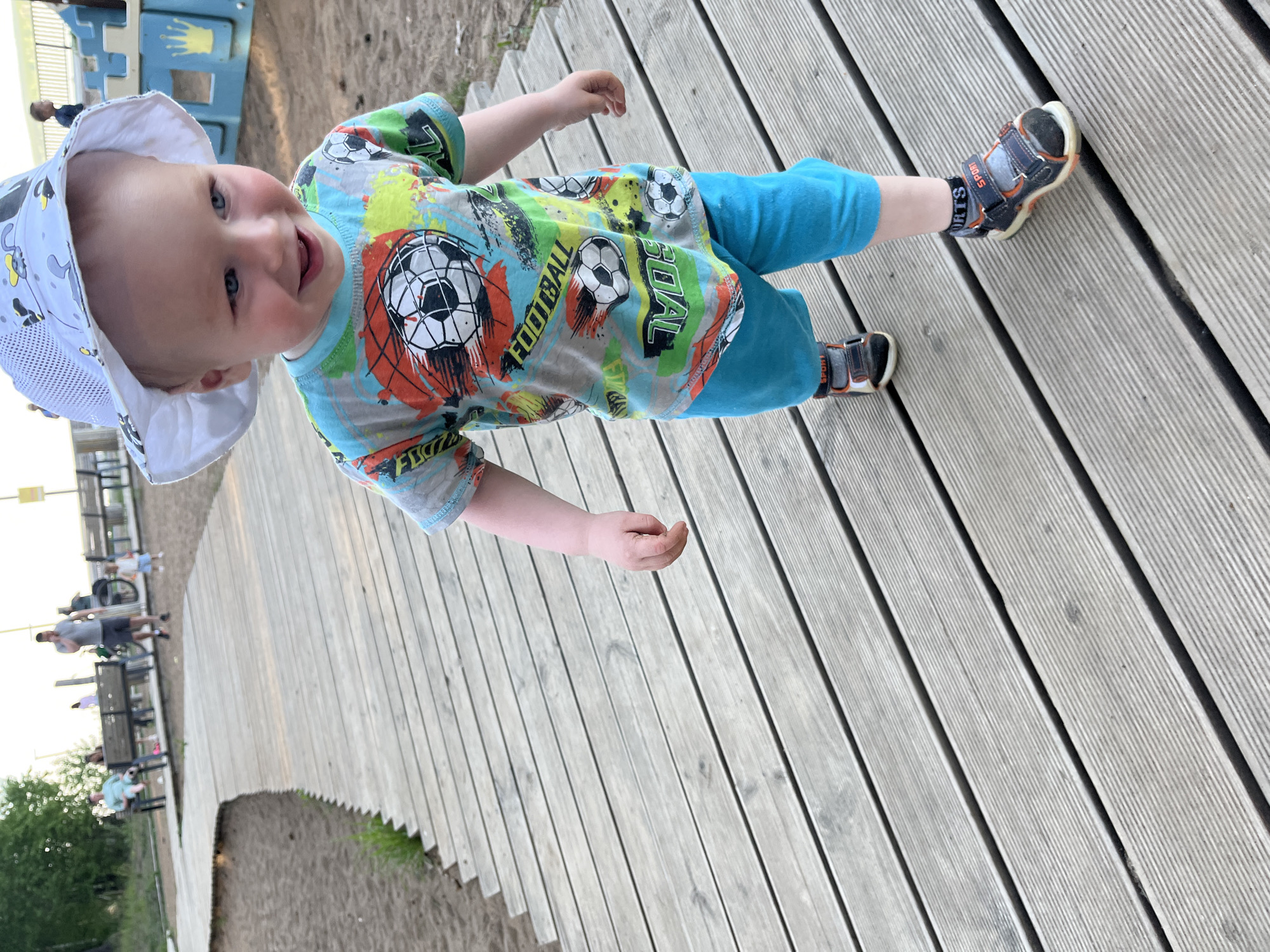 A toddler wearing a white sun hat, a soccer-themed t-shirt, blue pants, and sandals stands on a wooden boardwalk. The location appears to be a public park in Chaikovsky, Russia, with benches and other people visible in the background. The toddler is facing the camera and smiling. The boardwalk is made of light-colored wooden planks with spaces between them.