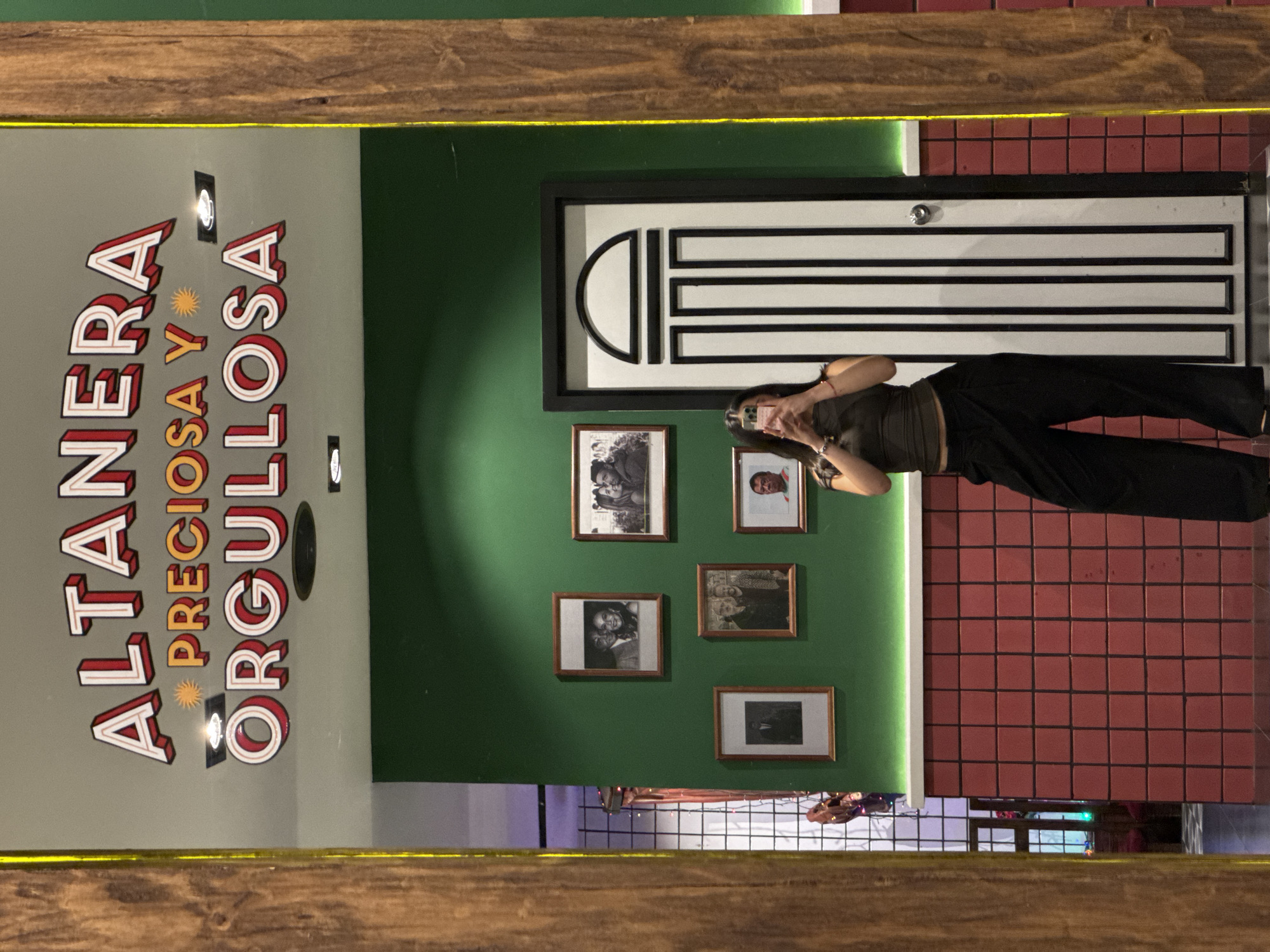 A female subject is captured in a mirror selfie within an interior space in Puebla, Mexico, identified by the slogan "ALTANERA PRECIOSA Y ORGULLOSA" displayed prominently. The subject, positioned centrally, holds a smartphone with both hands at chest level, facing forward to capture her reflection. She is dressed in a dark, possibly olive green, short-sleeved top and black wide-leg trousers, with dark hair visible. Red string bracelets adorn her right wrist. The mirror's frame is wooden with a thin yellow light strip integrated along its interior edge. The reflected background wall features distinct sections: an upper light greyish-green panel bearing the aforementioned slogan in stylized white, red, and orange lettering; a central dark green section displaying five framed black and white photographs of individuals and couples; and a lower section covered in reddish-brown square tiles, separated by a white horizontal ledge. A white door with a black outline, vertical black stripes, and a rounded top design is visible on the right side of the dark green wall, equipped with a chrome doorknob. Recessed lighting fixtures are visible on the upper wall panel.
