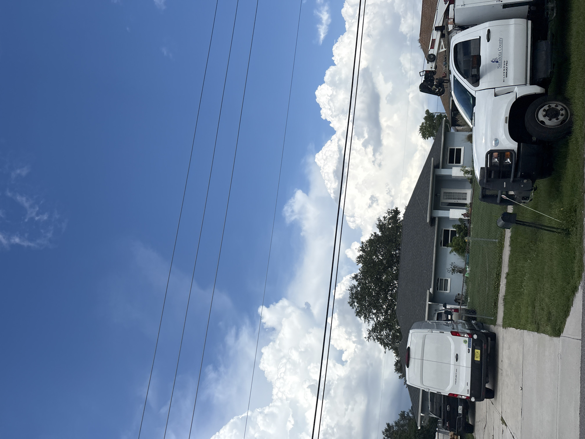 The image depicts a residential street scene in North Port, Florida, USA. In the foreground, a white utility truck with a crane attached is parked on the grass. The truck has "Sarasota County" and "Silverado 6600 HD" written on its side. To the left of the truck, a white cargo van is parked on a concrete driveway. Behind the van, a black pickup truck is partially visible. The background features a light blue single-story house with a dark roof, surrounded by a green lawn and trees. Power lines are strung across the sky, which is predominantly blue with large, fluffy white clouds. The overall impression is a suburban setting on a bright day, possibly with ongoing work or services taking place due to the presence of the utility vehicles.