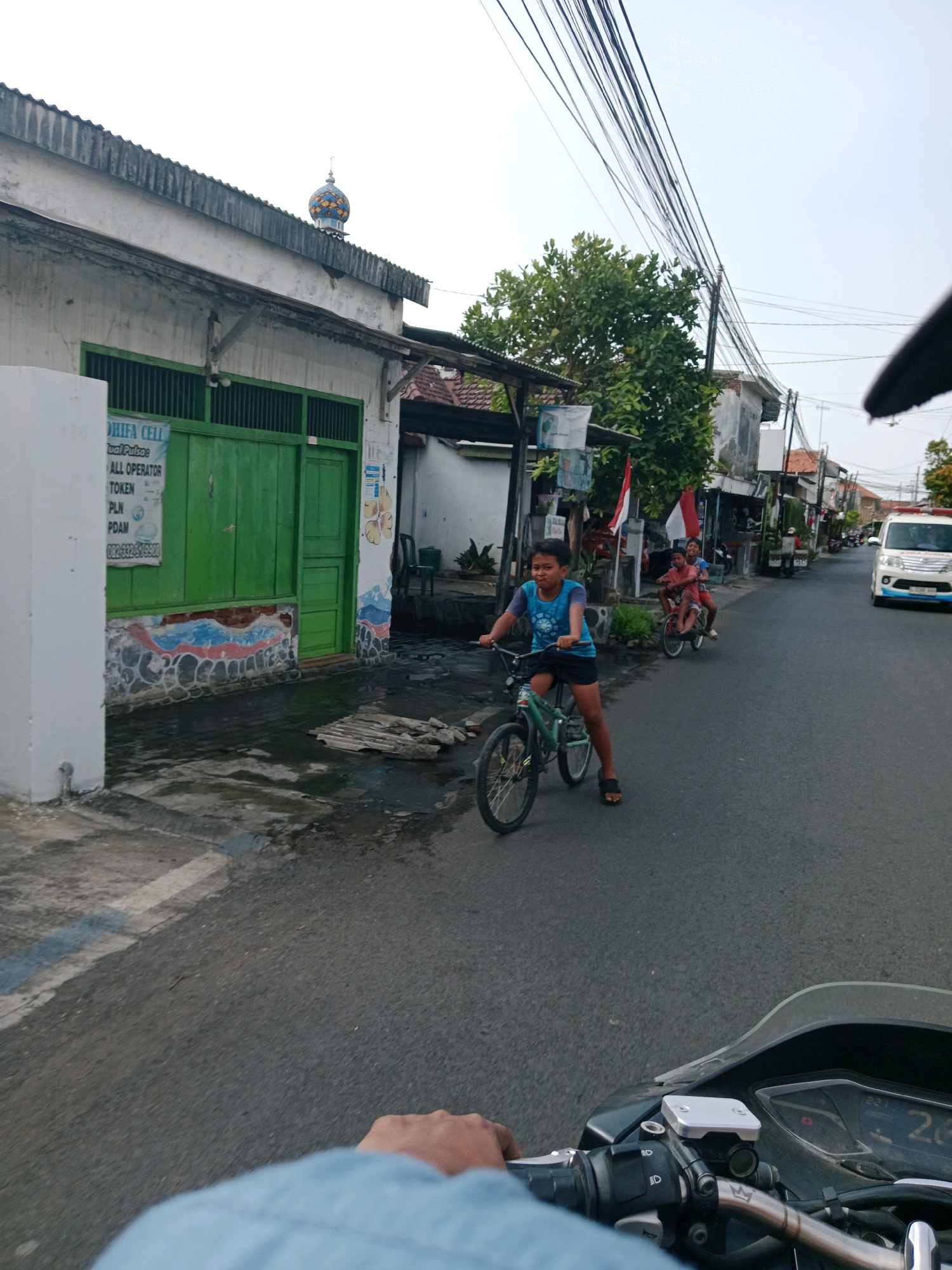 In Probolinggo, Indonesia, two young boys are captured riding bicycles on a paved street. The boy in the foreground, wearing a blue t-shirt and black shorts, is actively pedaling his green bicycle, his face contorted with effort. He wears sandals on his feet. Behind him, another boy, dressed in a red shirt and dark shorts, is also cycling, with a second person behind him on the same bicycle, though their details are less clear. The street is lined with buildings, one of which has a vibrant green door and a partially visible sign reading "DHIFA CELL." A small, colorful dome, possibly from a mosque, is visible atop one of the buildings in the distance. The sky is overcast, and the environment appears to be a residential or commercial area with power lines strung overhead. The perspective suggests the photo was taken from another moving vehicle, likely a motorcycle, as indicated by the handlebars and dashboard visible in the bottom right corner. The road surface shows signs of wetness, suggesting recent rain.