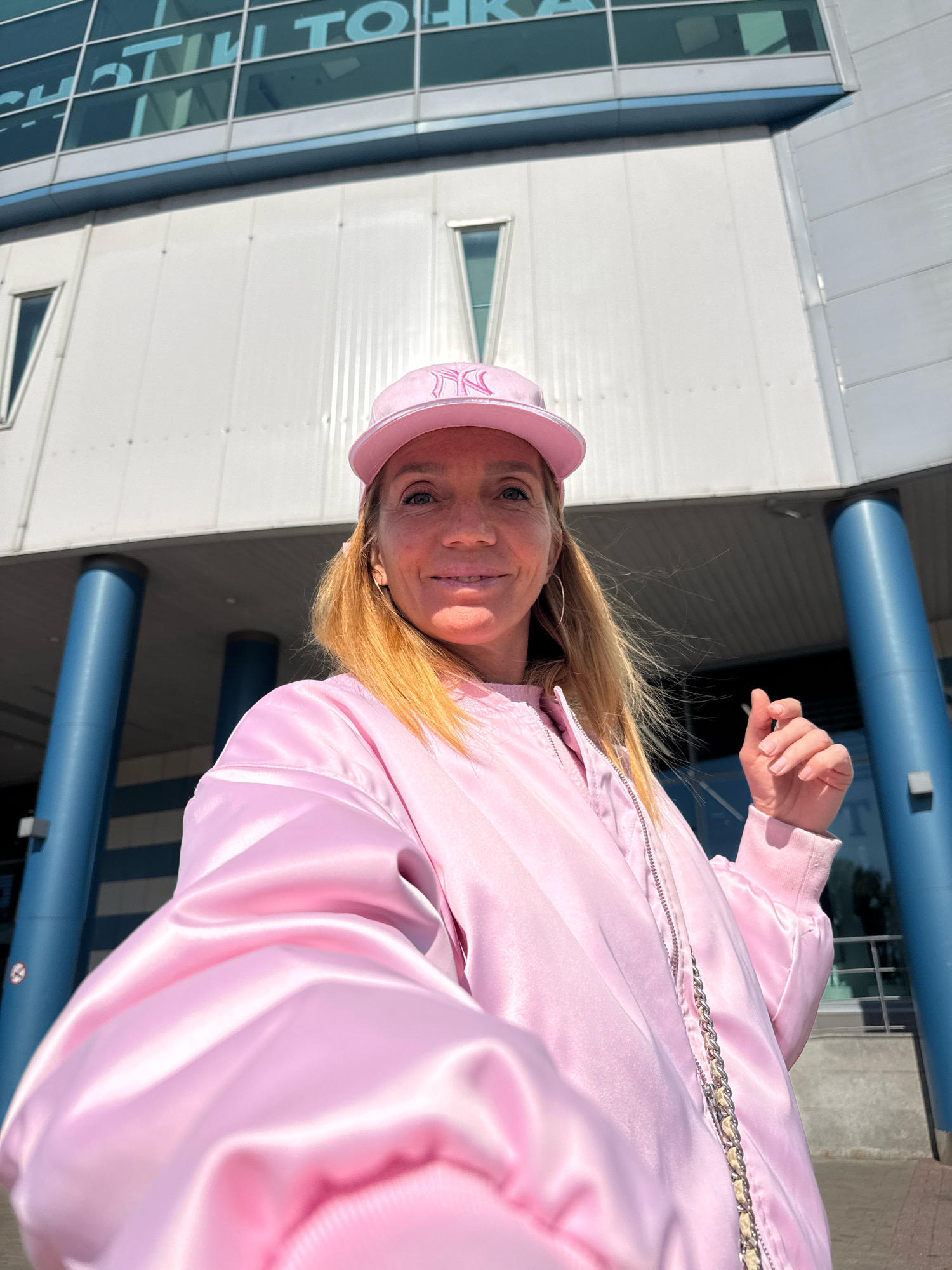 A woman with blonde hair wears a pink baseball cap and a pink satin bomber jacket with a chain detail. She is standing outside a modern building with blue columns and a glass facade. The building has text visible on its windows, including "STHIL TO..." and another segment of text which is illegible. The woman is smiling and looking at the camera, with her right hand raised slightly. The overall scene suggests a sunny day in a city environment.