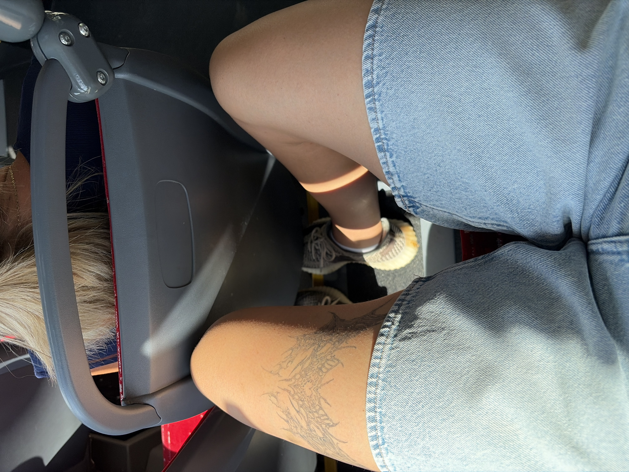 The media file shows a close-up view of two individuals seated next to each other on what appears to be public transportation, possibly a bus or train. The framing is from the waist down, focusing on the legs and clothing of the people.
One subject is wearing light-wash denim shorts and a tattoo is visible on their thigh. The tattoo is a dark line drawing of an intricate, possibly abstract or skeletal design. The other subject, partially visible, is also wearing denim shorts. Both individuals are wearing casual sneakers.
The setting appears to be inside a vehicle with gray plastic seating and a red accent on the seat structure. The lighting suggests it is daytime, with sunlight casting distinct shadows, particularly on the legs of the person with the tattoo. The environment is mundane, indicative of everyday travel. There is no visible text in the media file. The overall mood is neutral, capturing a moment of passive transit.
