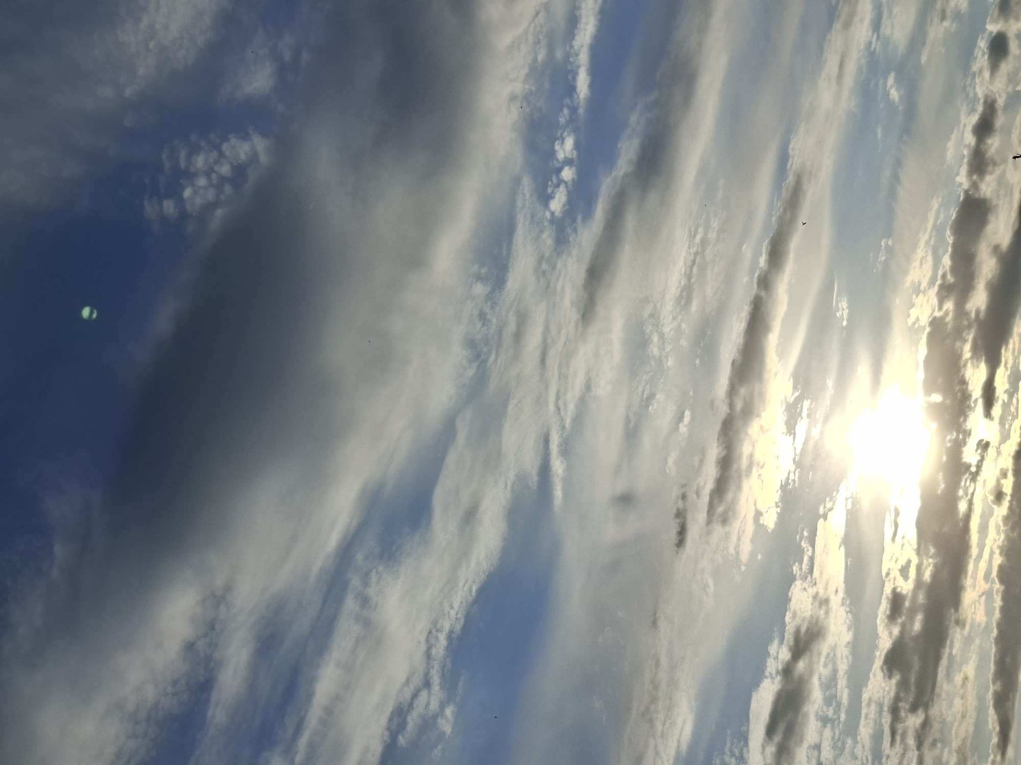 The image shows a sky with clouds and the sun. The clouds are a mix of dark and light shades, arranged in vertical streaks. The sun is bright and obscured by the clouds. There are a few dark spots in the sky, likely birds. The location is unknown.