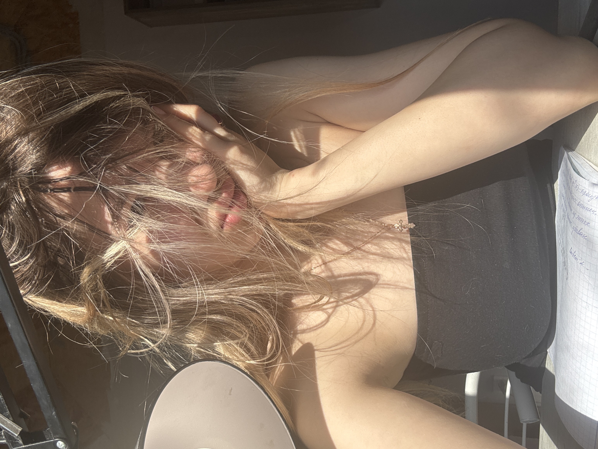 The image presents an angled, sunlit close-up of a person, likely female, with long, flowing blonde or light brown hair that partially obscures their face. Pink lips are visible amidst the hair and strong shadows. One arm is bent with a hand near the face, while the other arm rests across the body. A delicate gold-colored necklace featuring a small cross pendant adorns the person's neck.
The individual is wearing a dark, possibly black, strapless top or bandeau. The scene is illuminated by intense, direct sunlight, creating dramatic highlights on the hair and skin, and casting deep, intricate shadows across the subject and surroundings. This suggests a daytime setting where sunlight streams through a window or opening.
In the bottom right corner, a piece of white notebook paper with handwritten text in Cyrillic script is partially visible. Faintly discernible words include "читать" (read), "задач" (tasks/problems), and "задача" (task/problem), along with numbers like "2". To the bottom left, a circular, flat object, possibly a speaker or part of a lamp, rests on a dark surface. The background is generally dark and out of focus, with hints of furniture or room elements.
The overall mood is relaxed and contemplative, emphasized by the interplay of light and shadow. The visible Cyrillic text provides a subtle linguistic cue consistent with the location context of Noviy, Russia.