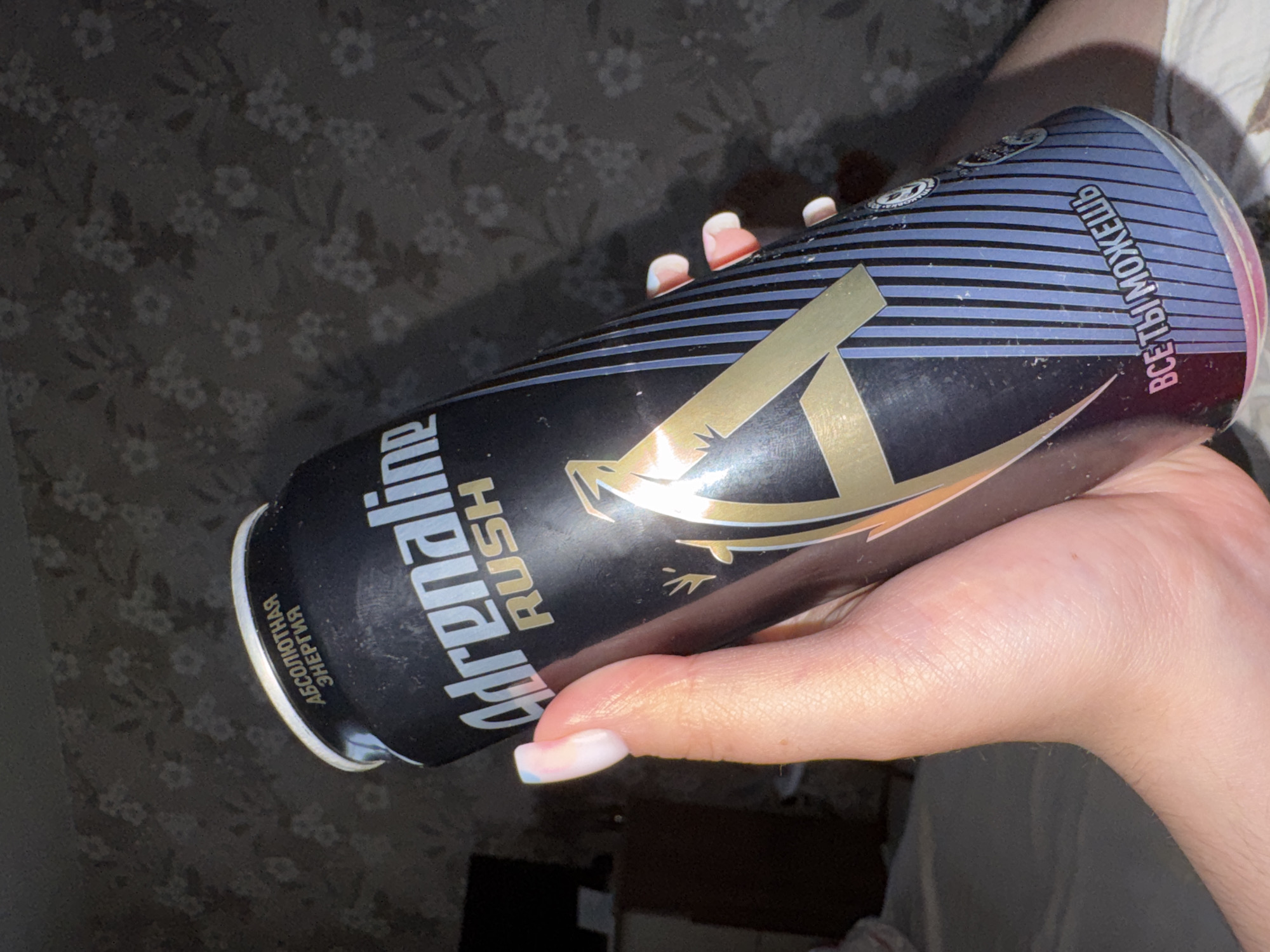 A close-up, angled shot features a light-skinned hand holding a can of "Adrenaline Rush" energy drink. The hand is positioned on the left side of the can, with the thumb and several fingers visible. The fingernails are manicured, appearing to have a light pink or white ombre design.
The energy drink can is primarily black with gold and blue accents. The prominent brand name "Adrenaline RUSH" is printed vertically in white and red text. Below "Adrenaline" and above "RUSH" in gold is a stylized letter "A" logo, which also incorporates the outline of a lizard or gecko. The can features horizontal blue stripes across its upper portion.
Several lines of Cyrillic text are visible on the can. On the left side, rotated, it reads: "АБСОЛЮТНАЯ ЭНЕРГИЯ" (Absolyutnaya Energiya), which translates to "Absolute Energy". On the right side, also rotated, it states: "ВСЁ ТЫ МОЖЕШЬ" (Vse Ty Mozhesh'), meaning "You Can Do Everything" or "You Can Do Anything".
The background consists of a dark grey or black fabric with a repeating, subtle floral or leaf pattern in a lighter grey, suggesting an indoor setting like a bed or sofa. The lighting is artificial, creating reflections on the metallic surface of the can. No specific activity beyond holding the can is depicted, and no particular time of day or emotions are discernible.