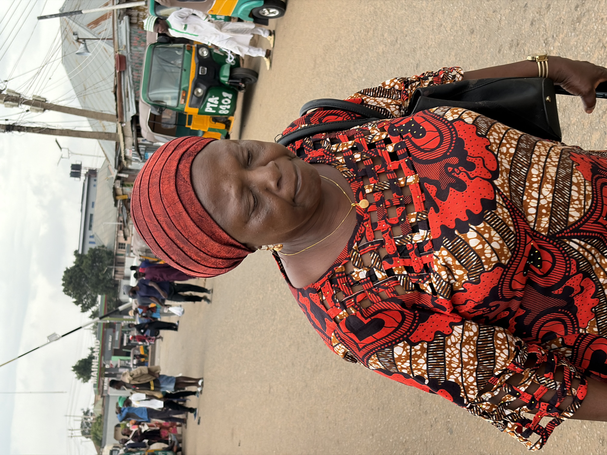 A woman wearing a red patterned headscarf and a matching patterned dress stands on a paved street in Gura Top, Nigeria. She is carrying a black handbag over her shoulder and wearing a gold bracelet and necklace. In the background, there are people walking on the street and a green and yellow vehicle. Overhead power lines are visible, and there is a gas station sign in the distance.