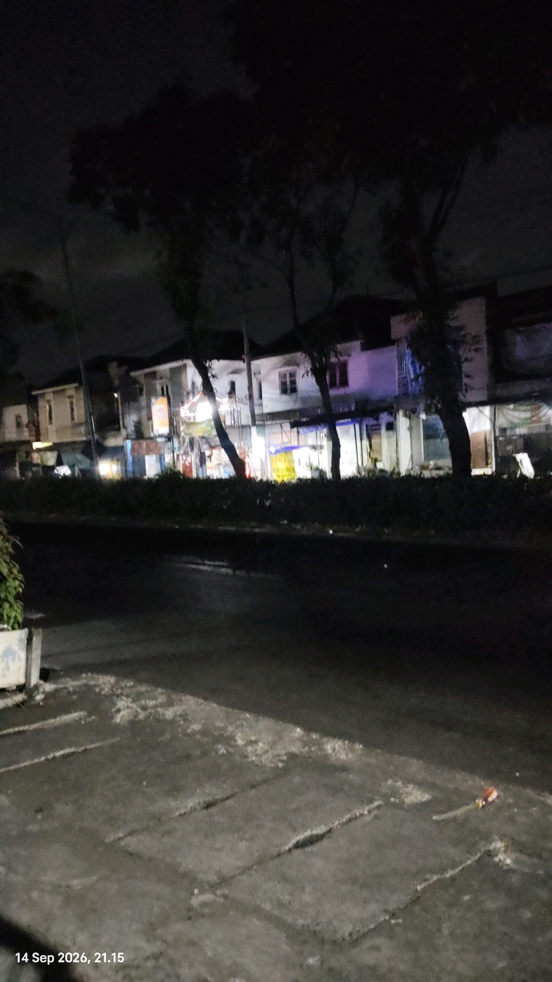 The image depicts a street scene at night in Surabaya, Indonesia. The foreground shows a paved walkway with visible concrete slabs and some scattered debris. A portion of a concrete planter box is visible on the left. The middle ground is a street with a dark, reflective surface, suggesting it may be wet. Beyond the street, a row of buildings is visible, illuminated by interior lights and streetlights. The buildings appear to be shops or businesses, with signs and awnings partially visible. Trees with dark foliage line the area between the street and the buildings. The sky is dark, indicating it is nighttime. A timestamp in the lower left corner reads "14 Sep 2026, 21.15".