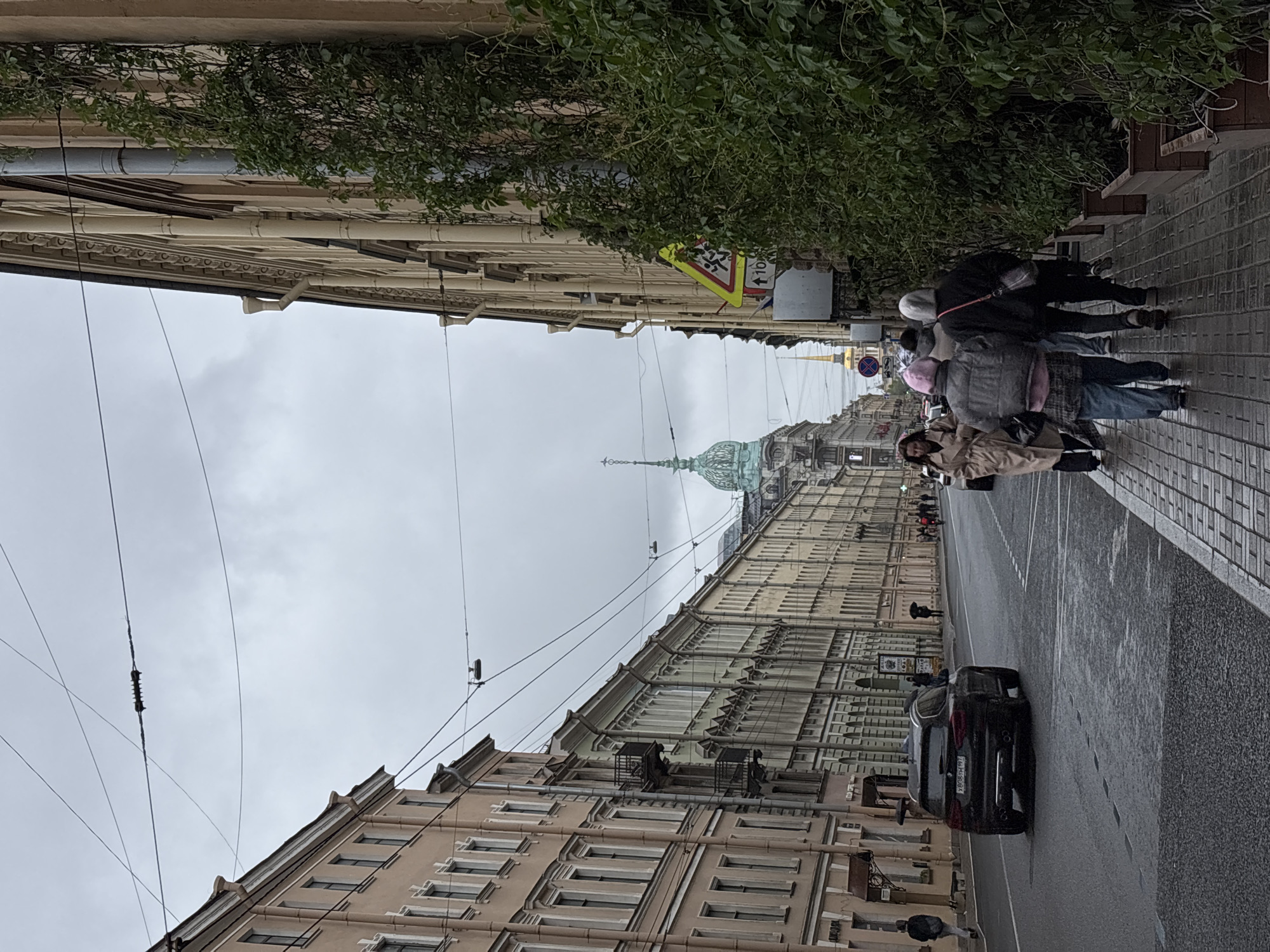 A slightly tilted eye-level shot shows a street in Saint Petersburg, Russia. On either side of the street stand tall, multi-story buildings with detailed facades, typical of the city's architecture. Above, a dull, overcast sky dominates the scene. Power lines crisscross overhead, adding to the urban landscape. The dome and spire of a major landmark building are visible in the distance, adding to the architectural interest of the image. On the right side of the frame, a group of pedestrians is walking along the sidewalk. They are bundled in coats and jackets, suggesting a cool temperature. The photograph captures the unique blend of historic architecture and modern urban life that characterizes Saint Petersburg.