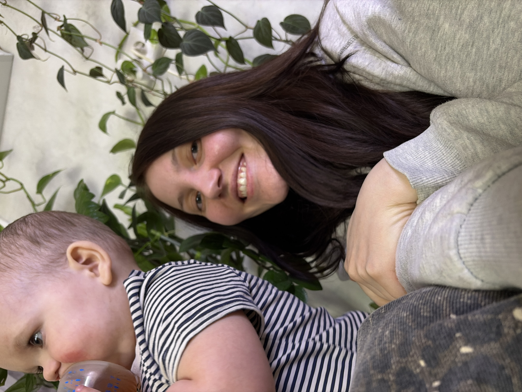 An indoor scene in Chaikovsky, Russia, depicts a baby and an adult woman. The baby, positioned on the left, is partially visible from the chest upwards, facing right with a slight upward gaze. The infant has fair skin, rosy cheeks, and light, short hair, and is wearing a navy blue and white horizontal striped short-sleeved top. The baby is actively drinking from a transparent plastic bottle adorned with blue polka dots, which contains an amber-colored liquid. A dark, possibly speckled fabric covers the baby's lower body, visible in the bottom left. To the right, an adult woman with long, dark brown hair smiles directly towards the viewer. She is wearing a light gray long-sleeved sweatshirt. Her left arm is bent, with her hand resting near the baby's leg, indicating proximity or support. The background consists of a plain light-colored wall adorned with several climbing plants featuring dark green leaves, trailing vertically and horizontally across the surface.
