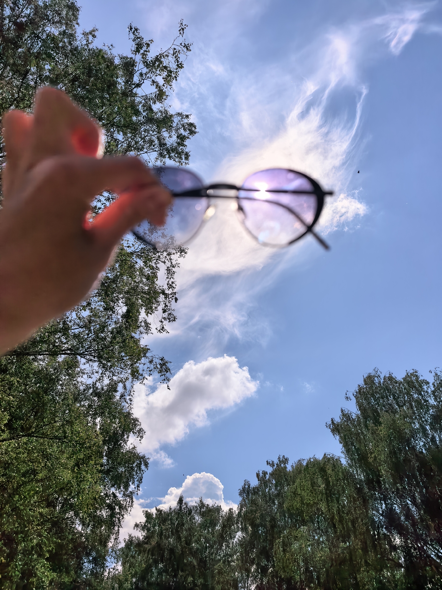 A hand holds a pair of sunglasses up to the sky. The sunglasses are round with a dark frame and tinted lenses that reflect a purplish hue from the sun, which is partially obscured by a wispy cloud. The background is a bright blue sky with scattered white clouds. Green trees with dense foliage frame the bottom and left sides of the image. The perspective is from below, looking upwards. The image does not contain any information that would indicate it is located in Sterlitamak, Russia, or any other specific city.