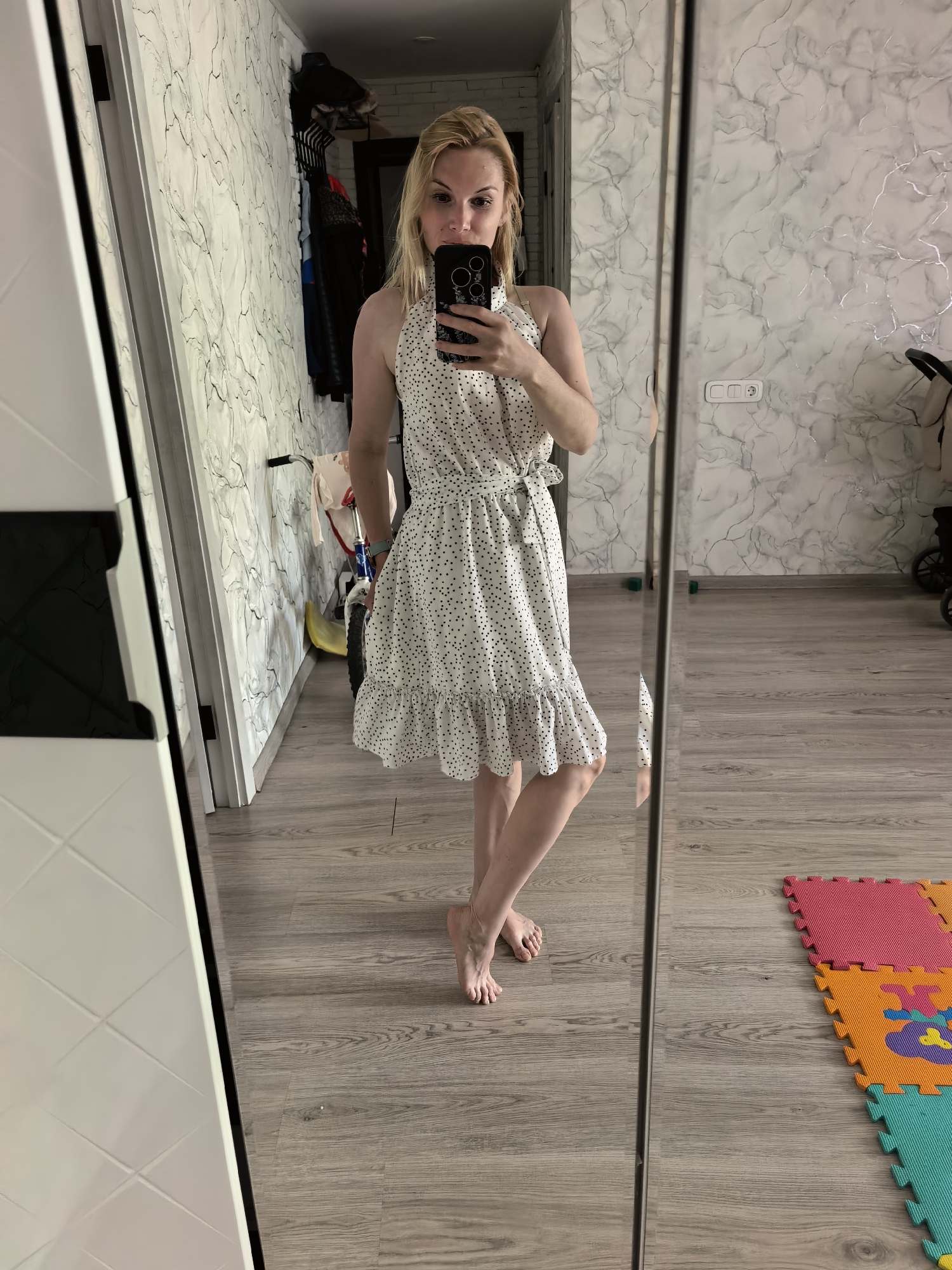 A woman is taking a selfie in front of a full-length mirror. She has blonde hair and is wearing a white halter dress with black polka dots and a tie at the waist. She is barefoot. The room has light wood-look flooring and white and gray marbled wallpaper. In the background are a baby stroller, a child's bicycle, and a clothes rack. The image shows sections of interlocking foam play mats in the lower right corner. The location is an apartment in Sterlitamak, Russia.