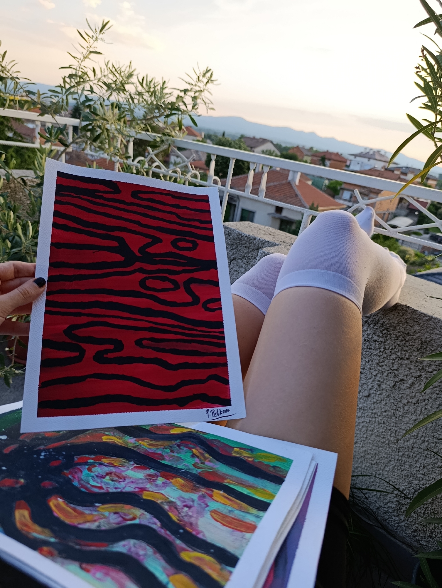 The image depicts a scene from a balcony or terrace in Svilengrad, Bulgaria, during daylight hours, likely approaching dusk or dawn. In the foreground, a person's bare legs are extended and rest on a textured, light grey concrete surface, clad in white knee-high socks. A left hand, with dark-painted fingernails, holds a rectangular painting vertically. This artwork features a red background traversed by irregular, thick black horizontal and wavy lines, reminiscent of wood grain or an abstract animal print. A signature, partially legible as "P. Petkov," is visible in the bottom right corner of this painting. Beneath the held painting, a stack of other artworks is present, with the topmost painting exhibiting abstract, wavy brushstrokes in vibrant colors including green, blue, yellow, red, and black.
The midground features a white, ornate metal railing, behind which various green plants are visible; some possess slender leaves, while others have broader foliage. Beyond the vegetation and railing, a residential area of Svilengrad unfolds, characterized by numerous buildings topped with orange-tiled roofs. In the background, a pale, hazy sky extends over a distant range of mountains or hills, completing the landscape view.