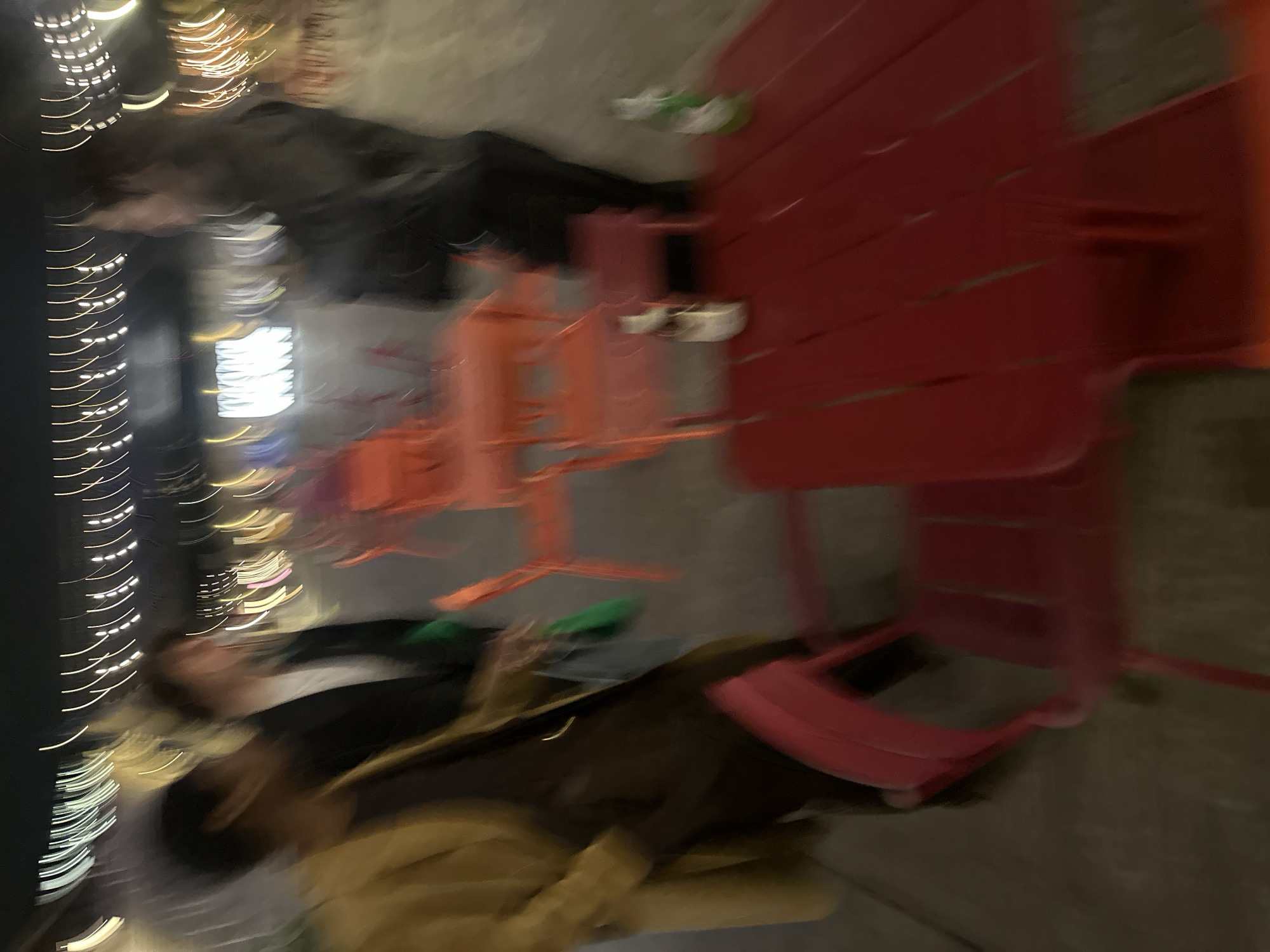 This blurred night-time image depicts an outdoor area in the city of London, United Kingdom, populated by multiple red and orange metal tables and chairs. In the immediate foreground, a red slatted metal table holds two bottles, one light-colored and one green, with a matching chair partially obscuring the table. Three individuals are visible, all heavily motion-blurred: on the far left, a person wears a yellow/gold coat over dark clothing; in the mid-ground, a person in a dark jacket and white top appears to hold a green object; and on the far right, a third individual in dark clothing stands near the tables. The background features linear streaks of artificial light, indicative of overhead fixtures in an urban pedestrian environment with a paved ground surface.