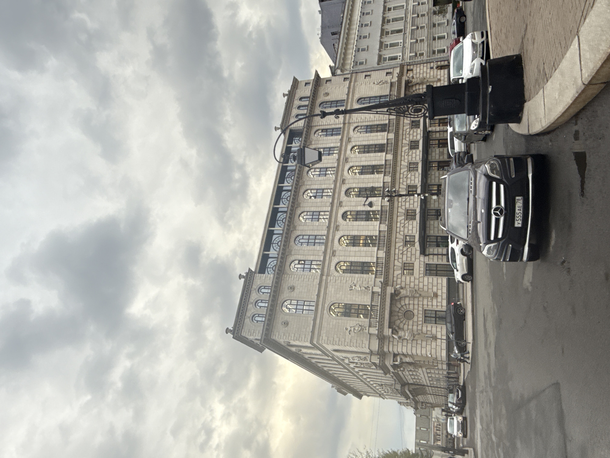 The image captures a street scene in Saint Petersburg, Russia, dominated by a large, ornate building with multiple arched windows and intricate architectural details. The sky is overcast, filled with gray clouds. A black Mercedes SUV is parked prominently on the right side of the street, with its license plate visible. Several other vehicles, including white vans and cars, are parked along the street. A lamppost with decorative wrought iron elements stands on the sidewalk. The street appears wet, reflecting light. No people are clearly visible in the immediate foreground. The building's facade suggests a historical or government building. The overall scene evokes a sense of urban ambiance under a somber sky.
