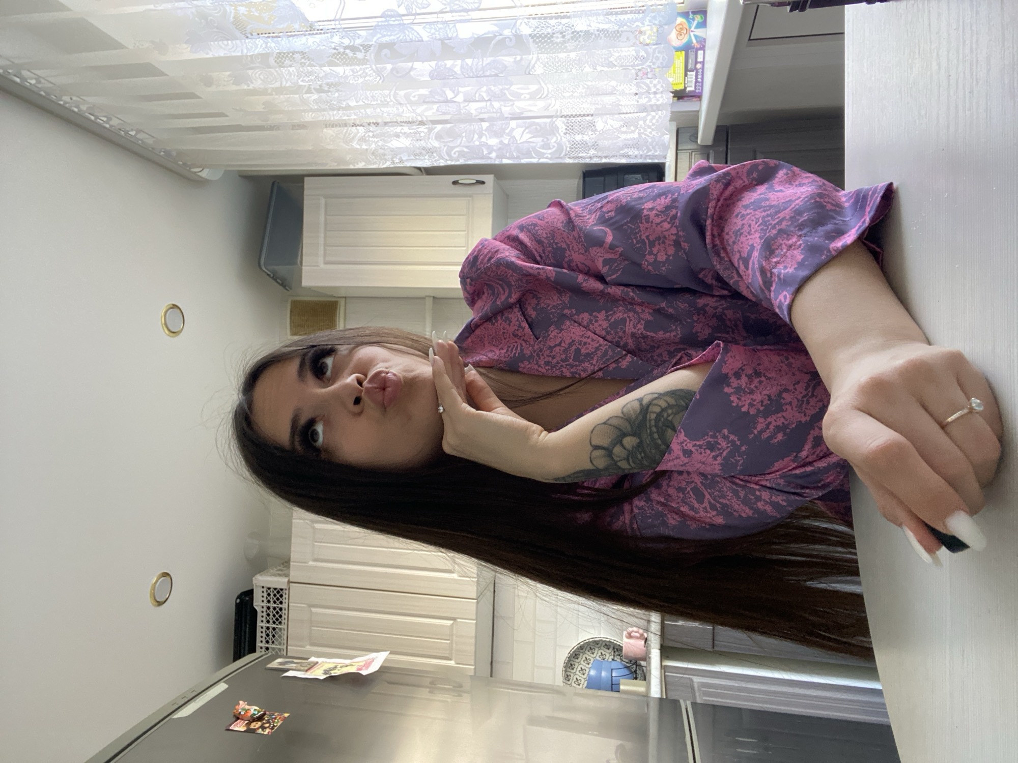 A young woman with long, dark hair is posing in what appears to be a kitchen. She is wearing a purple and pink patterned robe. Her right hand is touching her face, and she is pouting her lips towards the camera. She is wearing a ring on her right ring finger. Her left forearm has a large tattoo. The background consists of white kitchen cabinets, a white countertop, a refrigerator with magnets, and a window with white curtains. The exact location in Chaikovsky, Russia, is not discernible from the image alone.