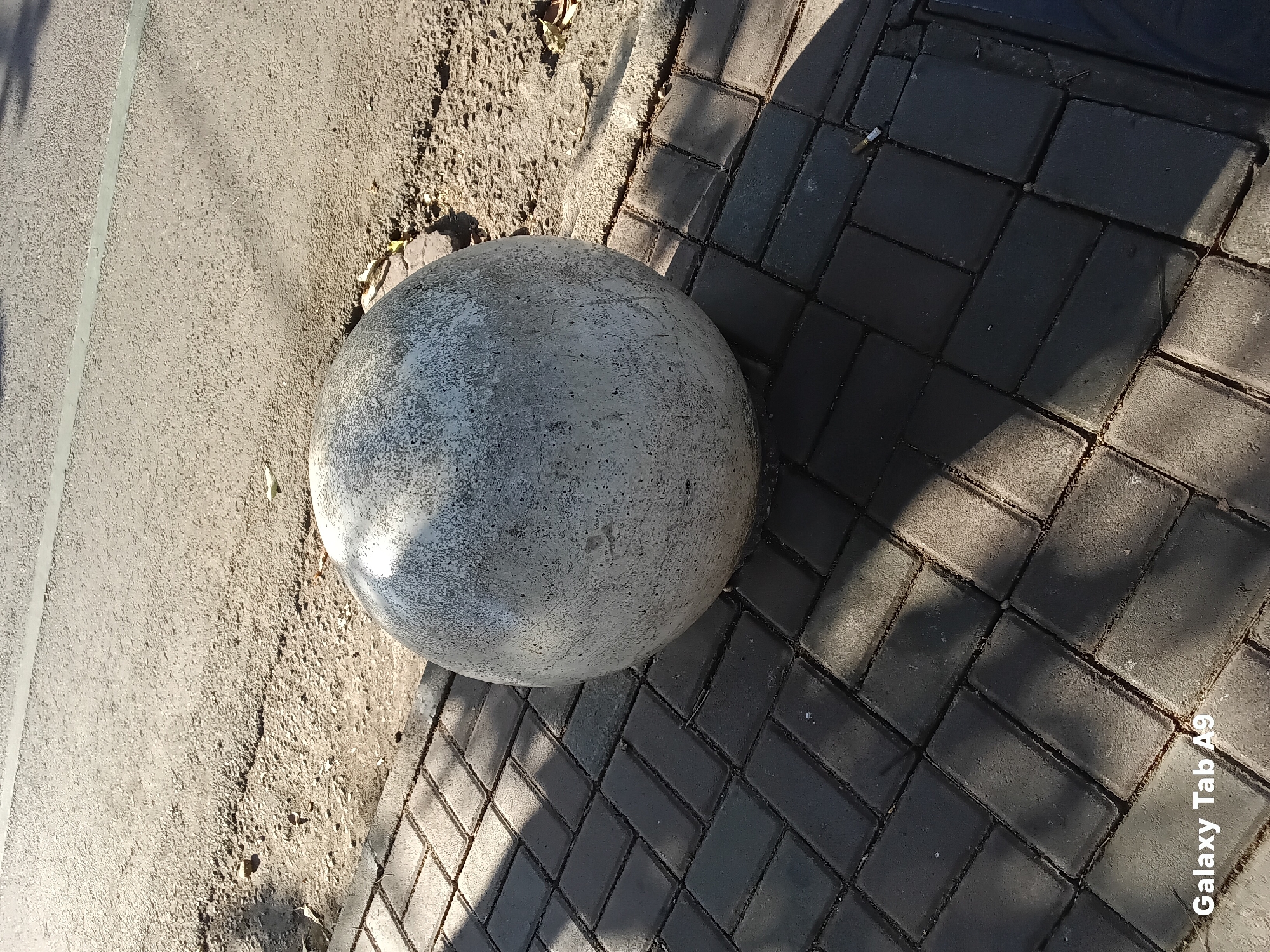 An outdoor, close-up, eye-level shot captures a large, spherical, weathered concrete bollard situated on a grey paver stone walkway adjacent to a paved road. The bollard, occupying the majority of the frame, exhibits a mottled texture with dark speckles against a lighter grey background. Sunlight casts a distinct shadow of the bollard onto the pavers, creating a diagonal line across the lower portion of the image. The road surface, to the right of the bollard, appears to be asphalt with a fine gravel texture and a subtle horizontal marking. The paver stones are rectangular and laid in a herringbone pattern, with visible grout lines. A few dry leaves and small pebbles are scattered on the ground near the base of the bollard. The bottom left corner of the image displays the watermark "Galaxy Tab A9". The scene is consistent with an urban or suburban street environment in Trenggalek, Indonesia, where such bollards are commonly used as traffic or pedestrian barriers.
