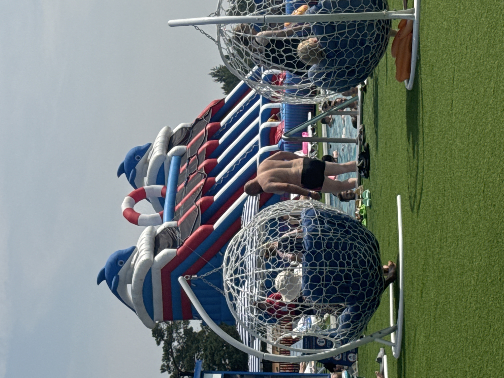 The image depicts an outdoor water park scene, likely in Votkinsk, Russia, given the context provided. In the background, a large, inflatable water slide with a dolphin theme is prominently featured, adorned in red, white, and blue. To the left and right of the slide are two large, white, spherical hanging chairs made of a mesh-like material, each containing several people. The ground in the foreground is covered in artificial green turf. In the middle ground, a man in black swim trunks stands partially in a swimming pool filled with people. The overall environment suggests a sunny day at a recreational facility.