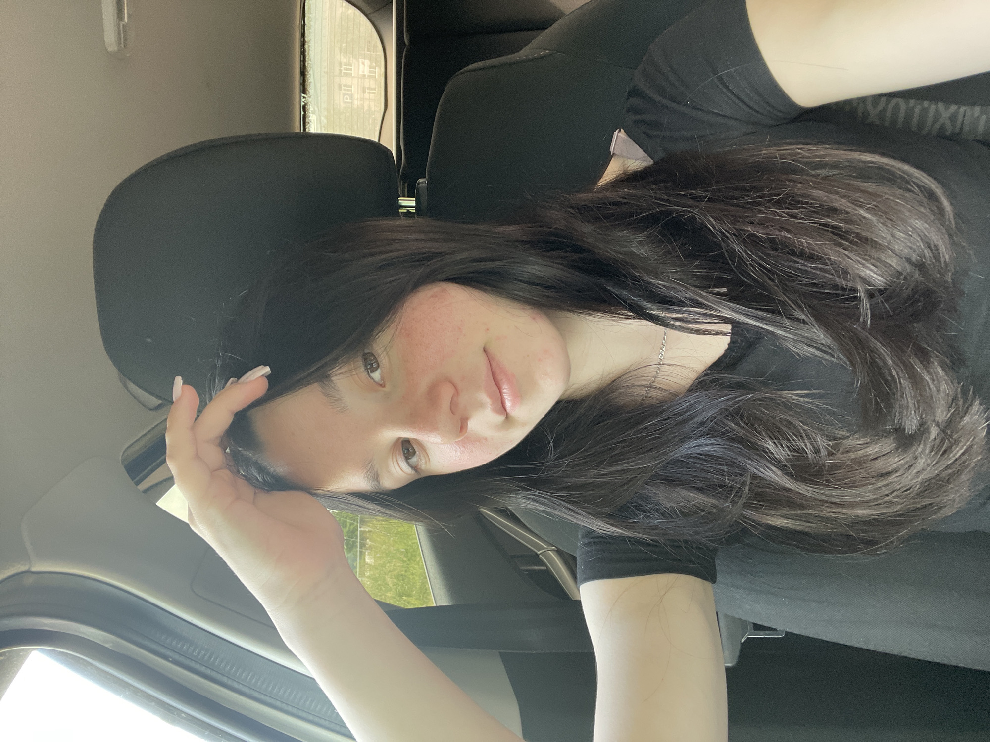 A young woman with dark hair and light skin is sitting in the passenger seat of a car. She is wearing a black t-shirt and a silver necklace. Her right hand is resting on her head, and her left arm is visible in the lower left corner of the image. The background shows the interior of the car, with a black headrest behind her. Outside the car, green vegetation and a fence are visible through the window. The image appears to be a selfie taken during daylight. The provided location, Nozhovka, Russia, is not verifiable from the image content.