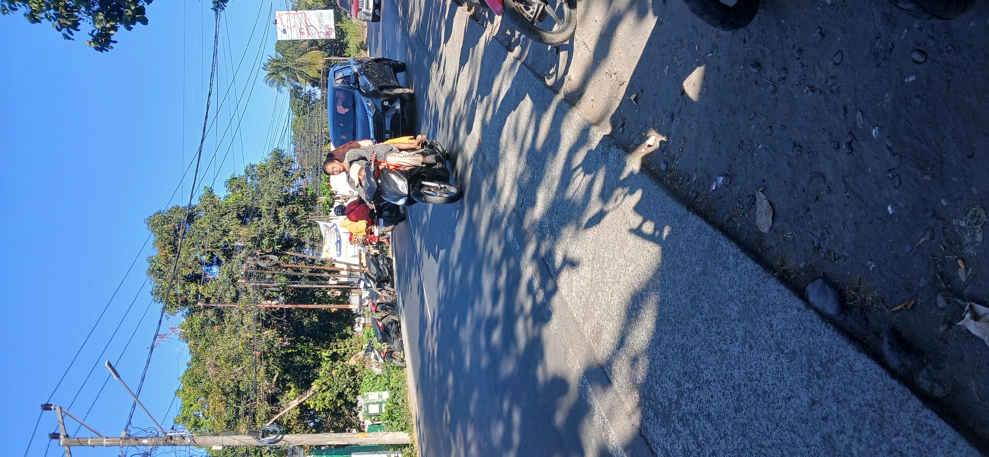 The image depicts a street scene in Kediri, Indonesia, under a clear blue sky. In the foreground, a dark-colored scooter is moving on a paved road, carrying two people. The driver, a woman, is wearing a light-colored top and patterned pants. A child, wearing a light-colored hooded garment, is seated in front of her. Behind the scooter, a dark-colored SUV is also on the road. To the left of the road, several parked motorcycles are visible, and a line of trees with dense green foliage forms the background. Electrical poles and wires are present overhead. The road surface is asphalt, with visible shadows of trees cast upon it. The right side of the road appears to be a dirt shoulder with scattered debris.