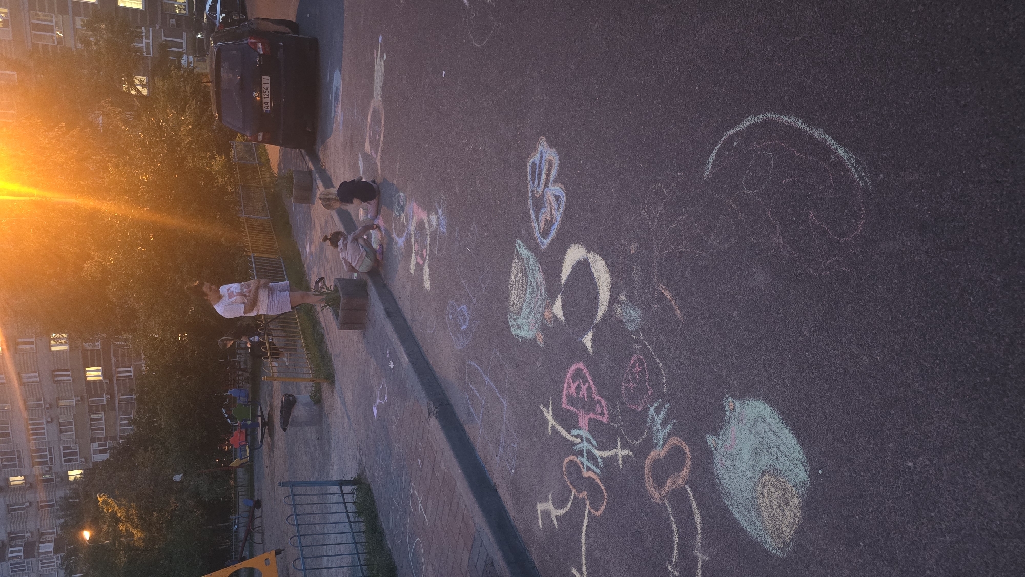 In Kyiv, Ukraine, a woman stands near a playground. Two young children are seated on the pavement, drawing with chalk. The pavement is adorned with various chalk drawings, including a blue and yellow drawing of a bird, a pink fish, and other abstract shapes and figures. In the background, a dark colored car is parked, displaying the license plate "AA 1254 II". Residential buildings and trees surround the area. The sun is setting, casting a warm orange glow over the scene.