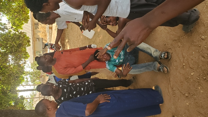 The image captures an outdoor scene in the city of Akwanga, Nigeria, showing a group of approximately ten adult males gathered on reddish-brown dirt ground under the canopy of several trees. In the immediate foreground on the right, a dark-skinned hand with a watch is visible, making a two-finger gesture.
Central to the composition, a male subject wearing a green Adidas t-shirt with "Standard Chartered" visible text and blue jeans is seated directly on the ground, his hands placed on his head, conveying distress. Partially obscured behind him, another male is seated or crouched, shirtless, also with his hands on his head in a similar posture.
Standing around these two individuals are multiple other males. From the left, one individual is seen in a dark blue, long flowing garment. Adjacent, a male wears a black and white patterned short-sleeved shirt and dark trousers. To the right of these, a prominent male wears an orange-red traditional long-sleeved garment and dark trousers. Further right, a male in a white short-sleeved shirt holds a white plastic bottle in his left hand and extends his right arm, pointing towards the arm of the male in the orange-red garment. A male in a white tank top is partially visible on the far right.
The background reveals a light-colored building with a sloped, tiled roof and two visible windows. Dry, light-brown vegetation is situated behind this building, and a light-colored motor scooter is parked nearby. The scene is brightly lit by natural daylight.