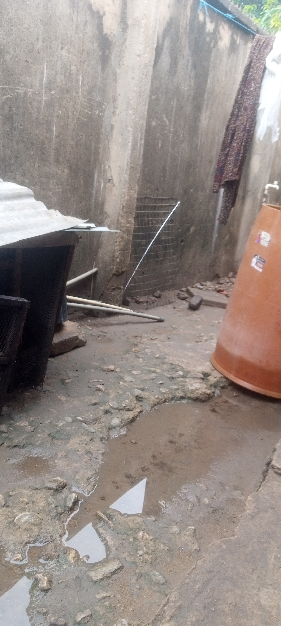 The scene is an outdoor space in Sabon Tasha, Nigeria. The ground is uneven and damp, with a puddle in the foreground reflecting the sky. A corrugated metal structure with a dark, enclosed area underneath is on the left. Two pipes lay on the ground near it. A wire mesh covers a section of the wall in the background. An orange container sits on the right. Clothes hang from a line on the wall further back. The walls are concrete, weathered, and stained. The overall impression is one of disrepair and dampness.