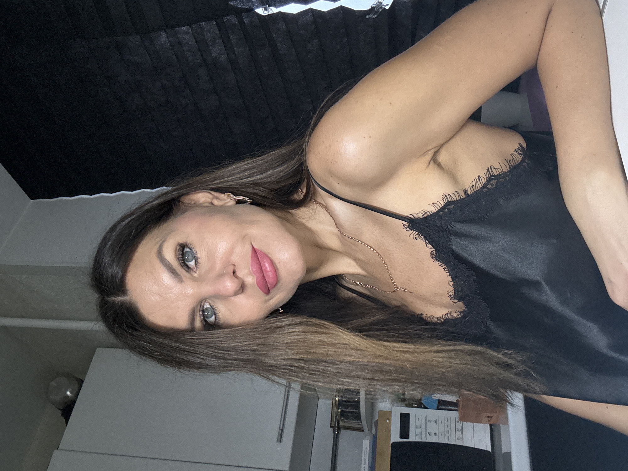 A woman is pictured indoors, likely in a home setting. She has long, brown hair and is wearing a black, lace-trimmed top with thin straps. She is adorned with earrings and a necklace. Her lips are colored with a pink hue. The background consists of a black curtain and a kitchen area with white cabinets and appliances. The photo does not contain sufficient information to confirm that the picture was taken in Chaikovsky, Russia.
