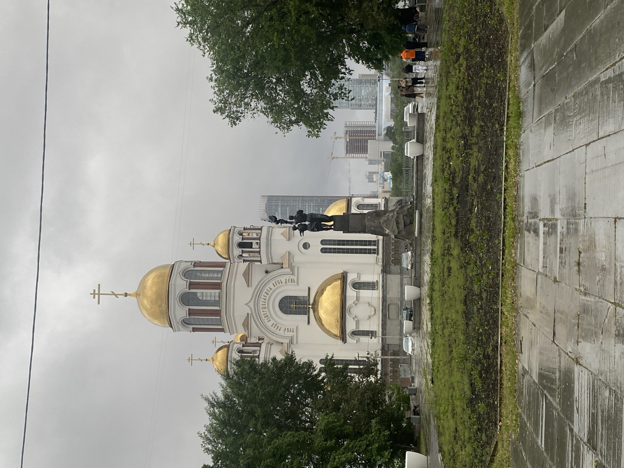 The image captures the Church on Blood in Yekaterinburg, Russia, a prominent landmark. The church is distinguished by its white facade and golden domes. The sky is overcast, indicating a gray or rainy day. In the background are modern buildings under construction, with cranes visible amidst the city's skyline. A pedestrian walkway, wet from a recent rain, runs alongside a green space. People are walking along the walkway in the background. A statue depicting several figures is located between the church and the walkway.