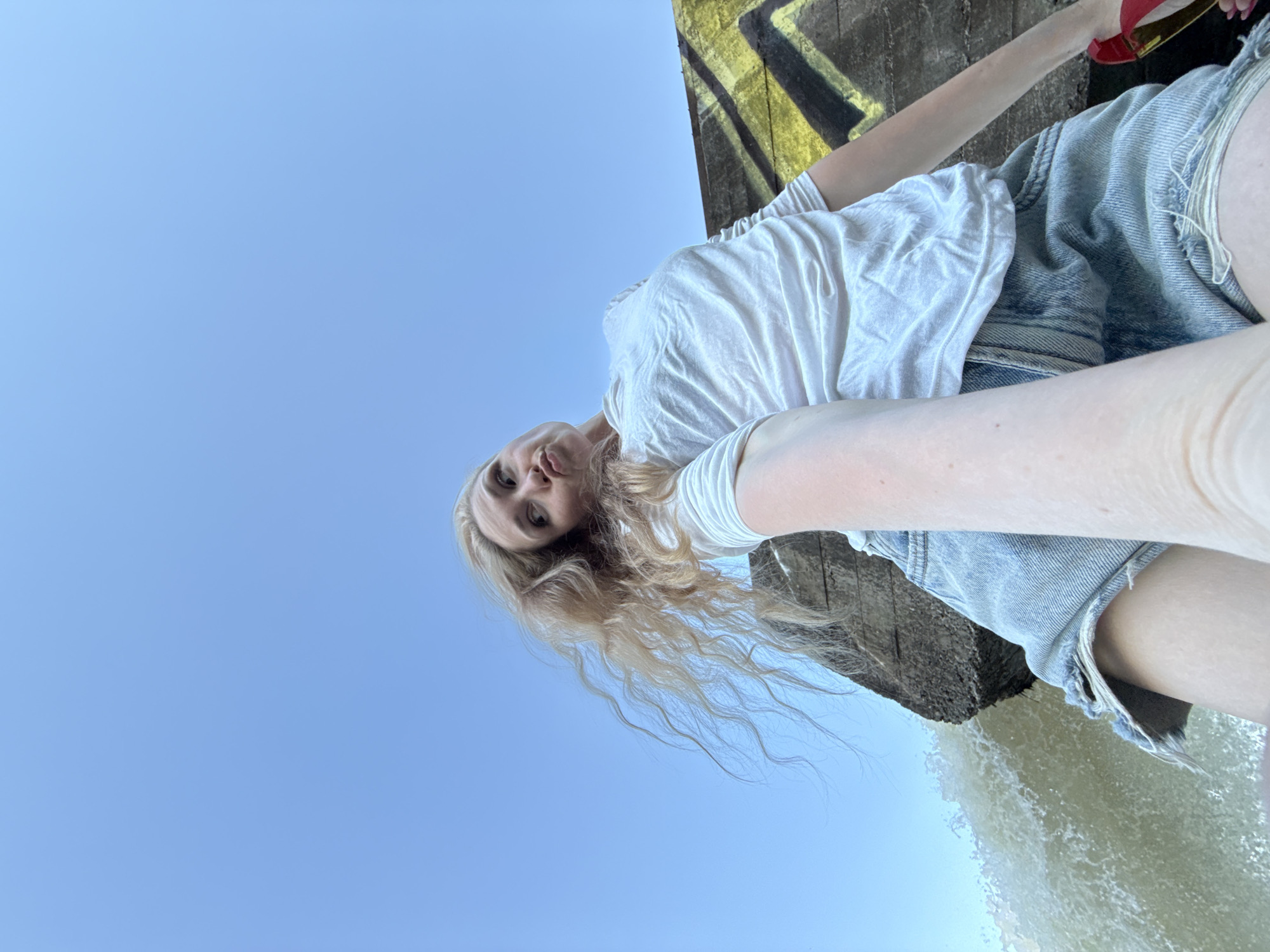 A young woman with blonde hair is positioned at the top of a concrete structure with graffiti on it, near what appears to be a waterfall. She is wearing a white t-shirt and denim shorts, and the background is primarily a clear blue sky. She is holding what appears to be a red cell phone in her right hand, as if taking a selfie. The location is Novyi, Russia.