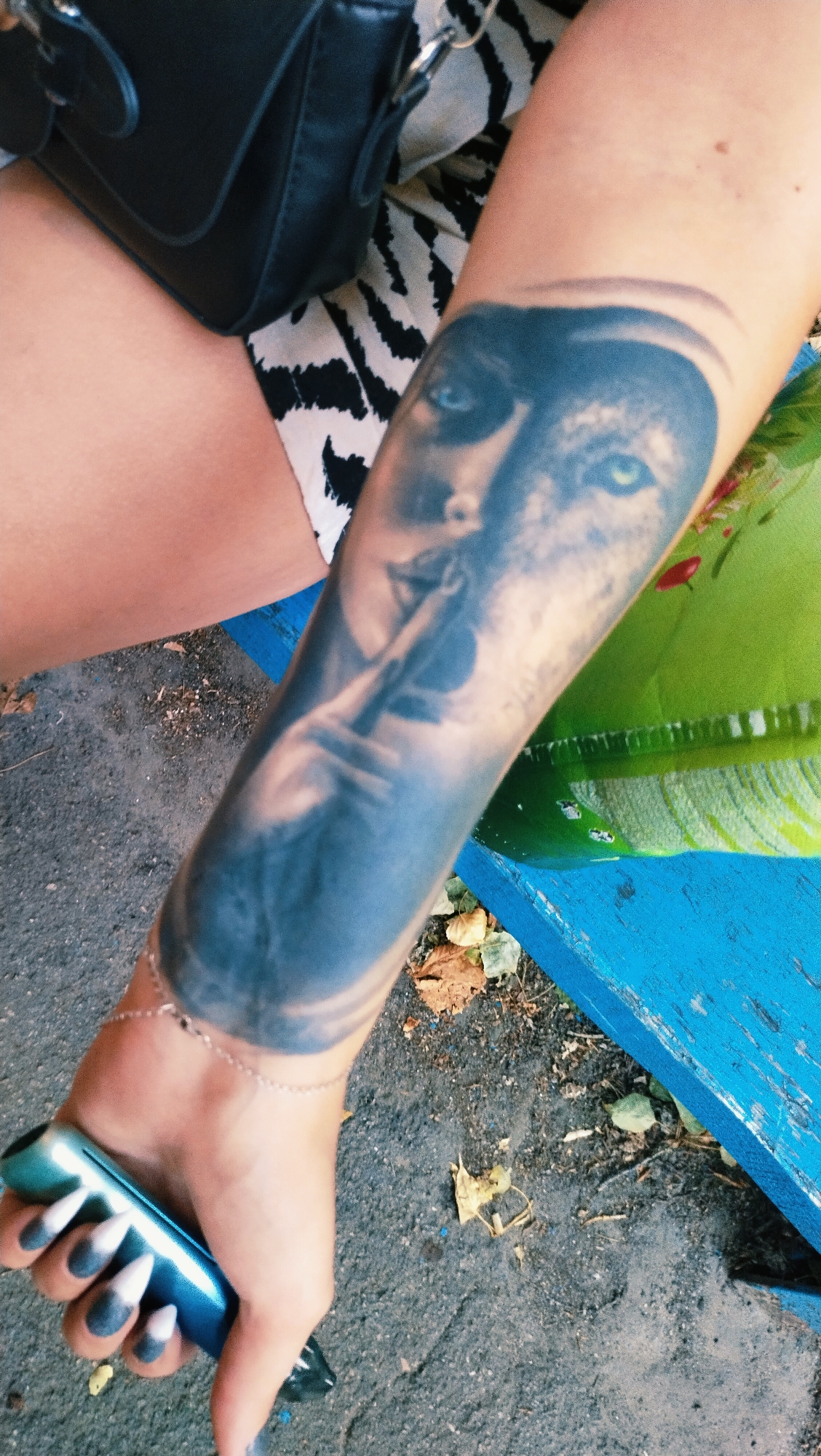 The image shows a close-up of a person's forearm with a tattoo of a face. The tattoo is a realistic depiction of a woman with bright blue eyes and her finger to her lips. The forearm also has a delicate silver bracelet with small black beads. The person's hand is holding a blue vaping device with a metallic sheen. The person's fingernails are painted in a dark grey and white gradient design.
The person is wearing shorts with a black and white zebra print pattern and a black handbag is visible in the background. The tattoo is on the person's right arm, and they are seated outdoors. The ground appears to be paved with asphalt or gravel, and fallen leaves are scattered around. A bright blue wooden surface, possibly a bench, is visible to the right of the forearm.
The image was taken in the city of Horishni Plavni, Ukraine.
