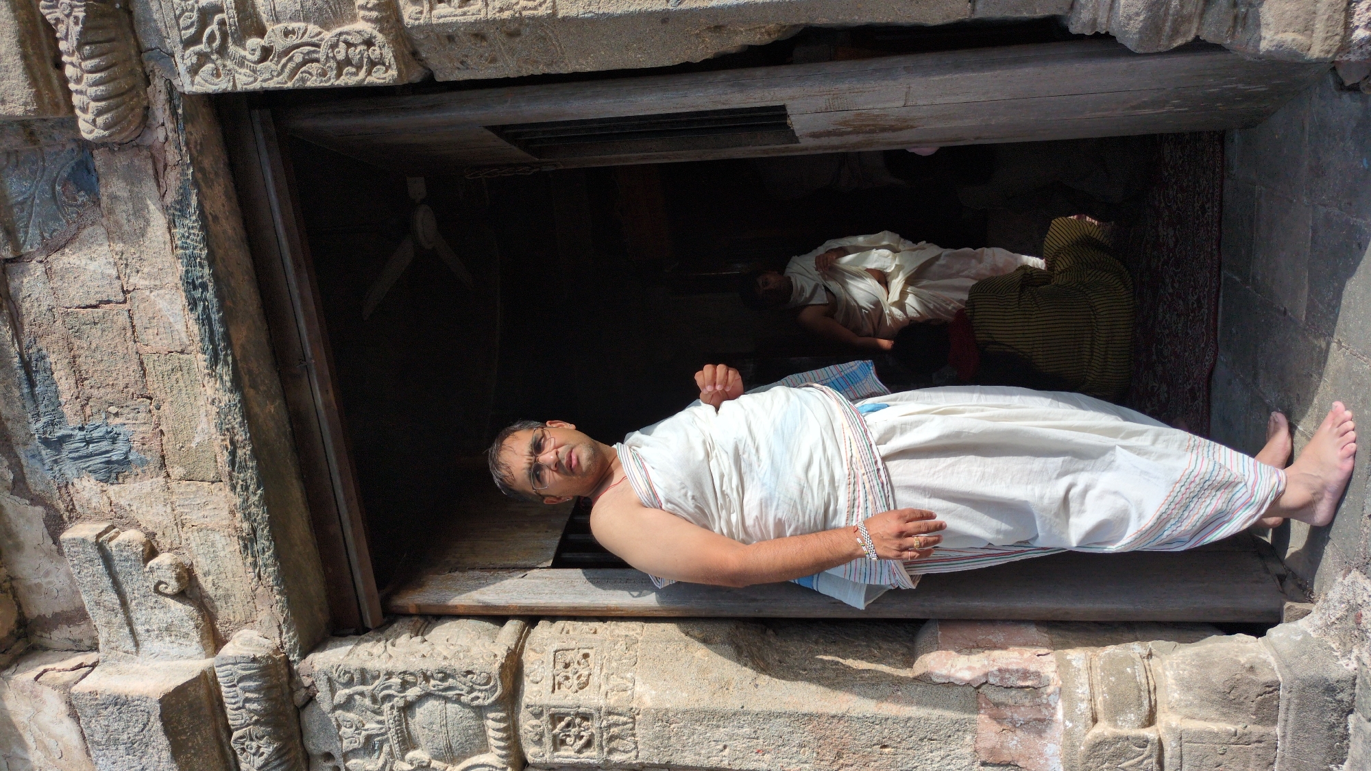 An angled, outdoor medium shot shows a man lying in a doorway or opening of a building made of chiseled stone, with others lying on the floor inside. The setting is in Kaswara, India. The man in the foreground is wearing glasses and is clad in a white cloth. He has a watch and ring on. His bare feet are at the right edge of the shot. Two other people are lying on the floor in the dimly lit interior, covered in white and yellow striped cloths, respectively. A ceiling fan is also visible inside. The stone exterior surrounding the doorway is ornately carved.