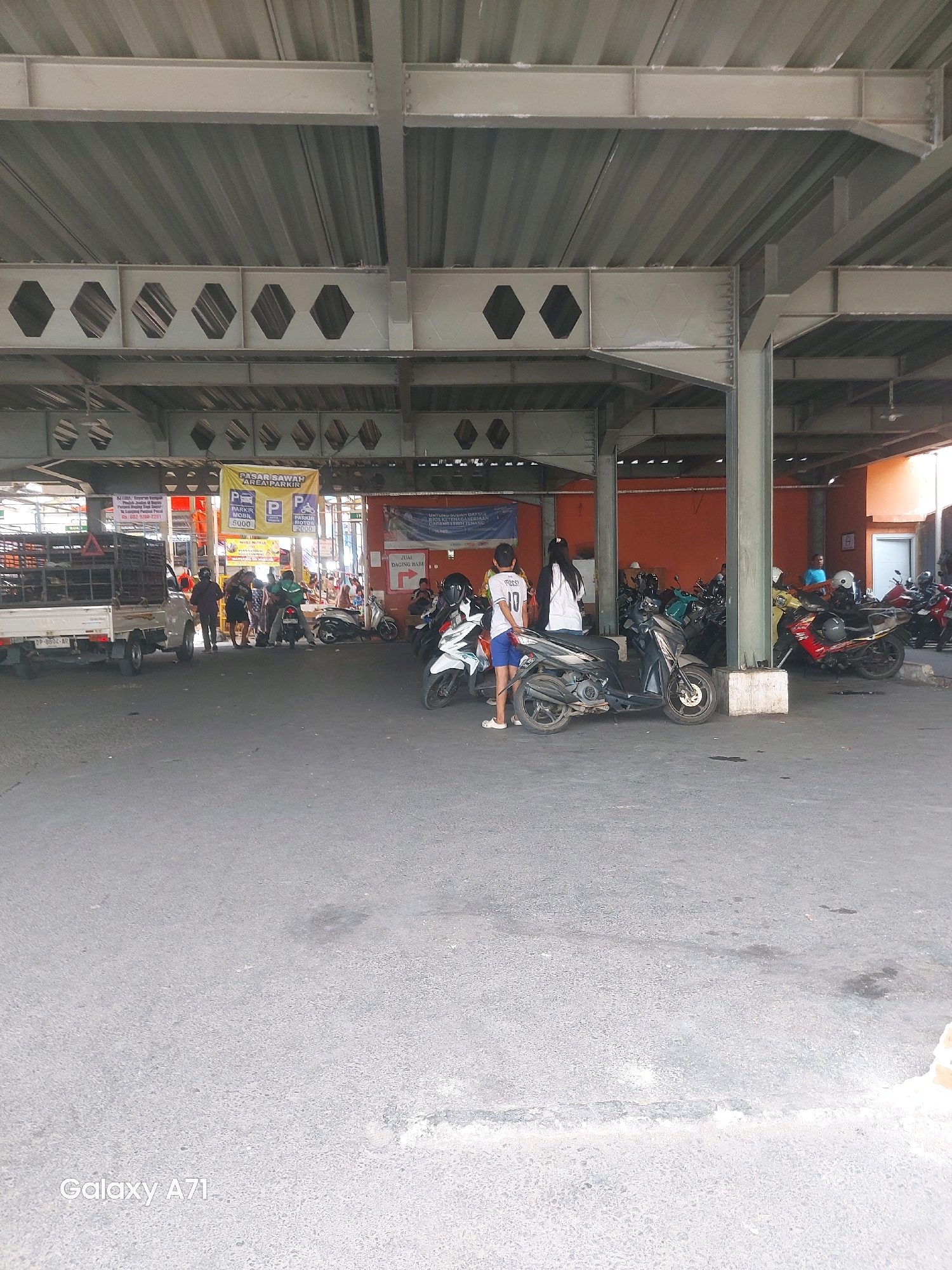 An outdoor parking area for motorcycles and some vehicles is depicted under a metal roof structure. The parking area appears to be part of a market or a public facility. Several motorcycles are parked in rows, with some individuals interacting near them. A white truck with a cage is parked to the left of the frame. In the background, there is an orange wall and a building. A yellow banner with "PASAR SAWAH AREA PARKIR" (Market Field Parking Area) is visible. The ground is paved asphalt. The image also shows a watermark "Galaxy A71" in the bottom left corner. The location is identified as Makassar, Indonesia.