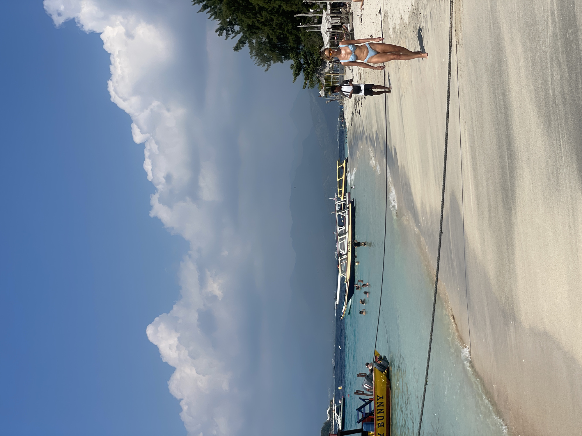 This image depicts a tropical beach scene in Unknown, Indonesia. The foreground shows a sandy beach with a rope barrier running horizontally across it. The turquoise water of the ocean is visible on the left, with small waves lapping the shore. Several boats are anchored or moored in the shallow water, including a bright yellow boat with "Y BUNNY" written on its side. In the distance, more boats and the hazy outlines of mountains are visible. The sky is a clear blue with scattered white clouds.
On the beach, a woman in a light blue bikini walks towards the right side of the frame. She is wearing sunglasses. Behind her, a man in a black t-shirt and shorts walks away from the camera. Further down the beach, there are people swimming and wading in the water, as well as some beach chairs and umbrellas set up under trees. The overall atmosphere is one of a relaxed beach vacation.