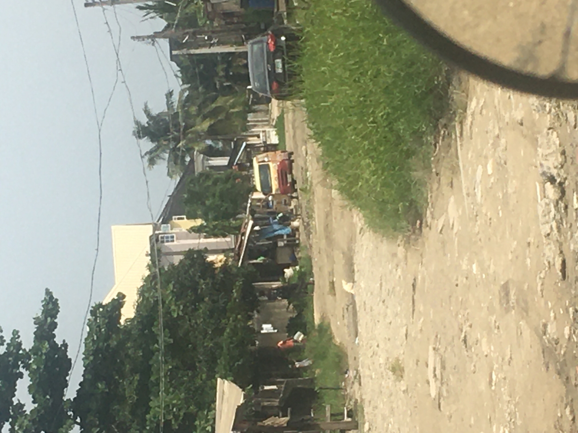 The scene depicts an unpaved road in Iranla, Nigeria. Along the roadside, there is a mix of vegetation and informal dwellings. A yellow and red vehicle is parked on the side of the road. Farther down the road, a dark-colored car is visible. A tall, light-colored building is partially visible behind trees. Utility poles with tangled wires are present. The environment appears to be impoverished with signs of disrepair and informal settlements.