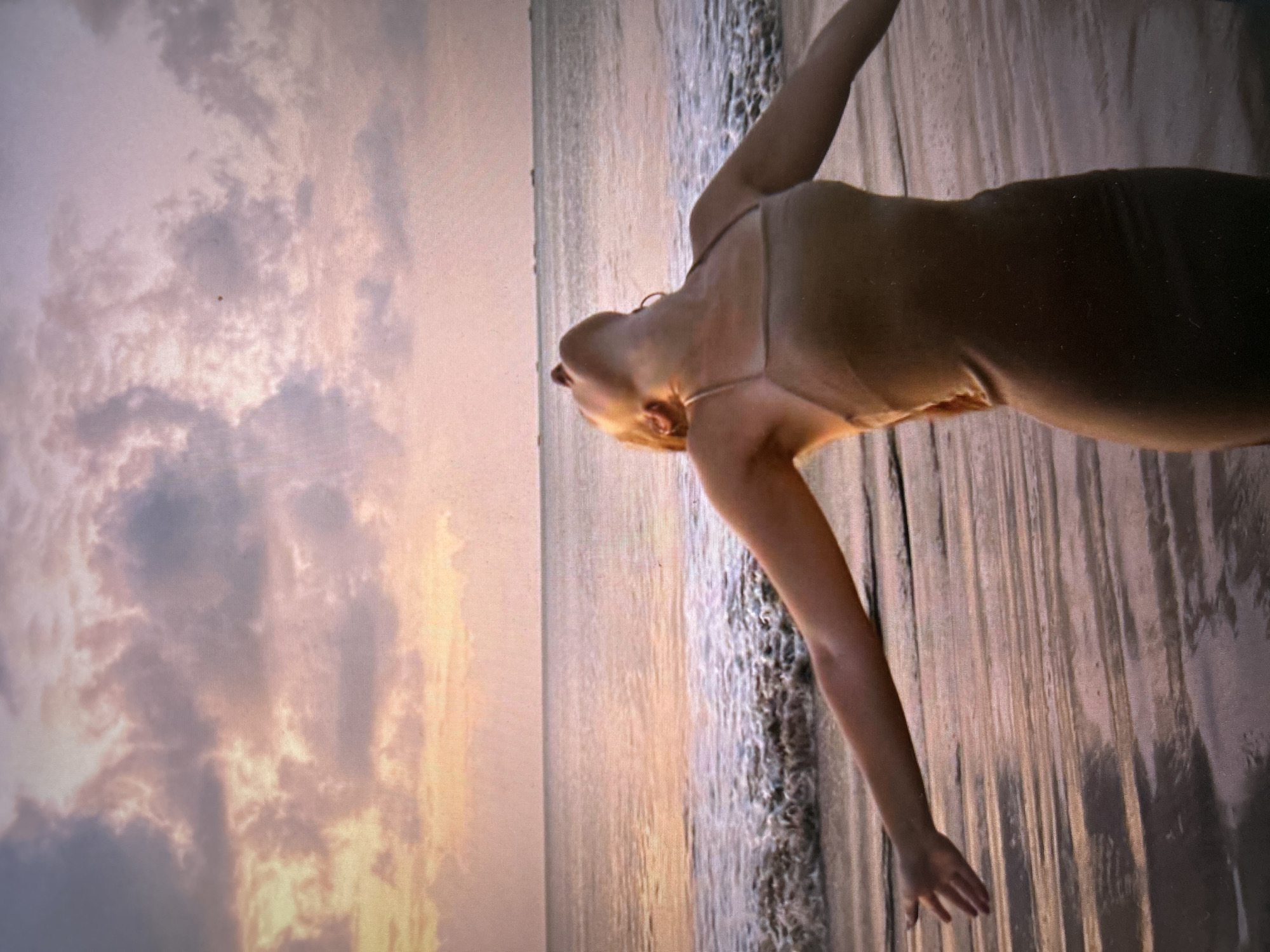The image captures a person in a dress, lying on the beach at sunset. The beach is likely in Kuta Selatan, Indonesia, given the typical scenery. The person is positioned with their body facing the ocean, head tilted back, and arms outstretched. Gentle waves are washing over the sand. The sky is filled with clouds illuminated by the setting sun, creating a warm, golden light. The photograph is taken at a low angle, emphasizing the sky and the person's pose.