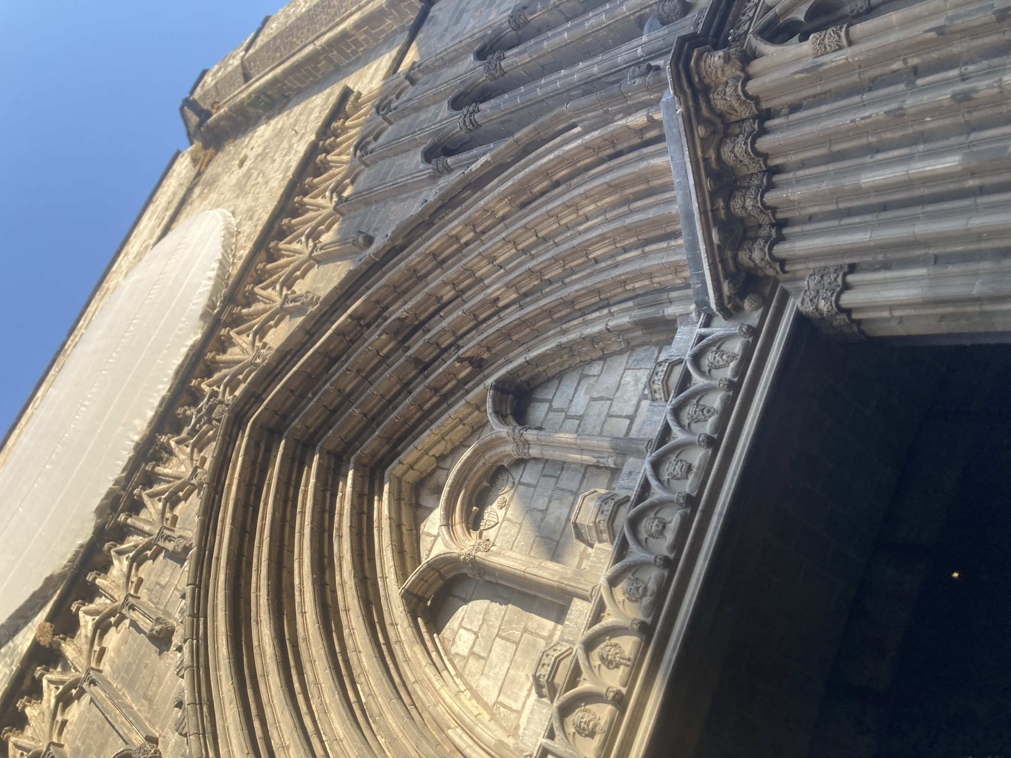 The image shows an ornate stone entryway of a Gothic cathedral in Barcelona, Spain. The detailed carvings include multiple arches, columns, and decorative elements such as faces and what appear to be architectural ornamentation. The stone is weathered and shows signs of age, with some areas appearing darker than others due to time and elements. Above the main archway, a large section of the building is covered by a white tarp, suggesting ongoing restoration or maintenance. The sky is a clear, bright blue, indicating a sunny day. The perspective is looking up at the cathedral from a low angle, emphasizing its grand scale and architectural complexity.