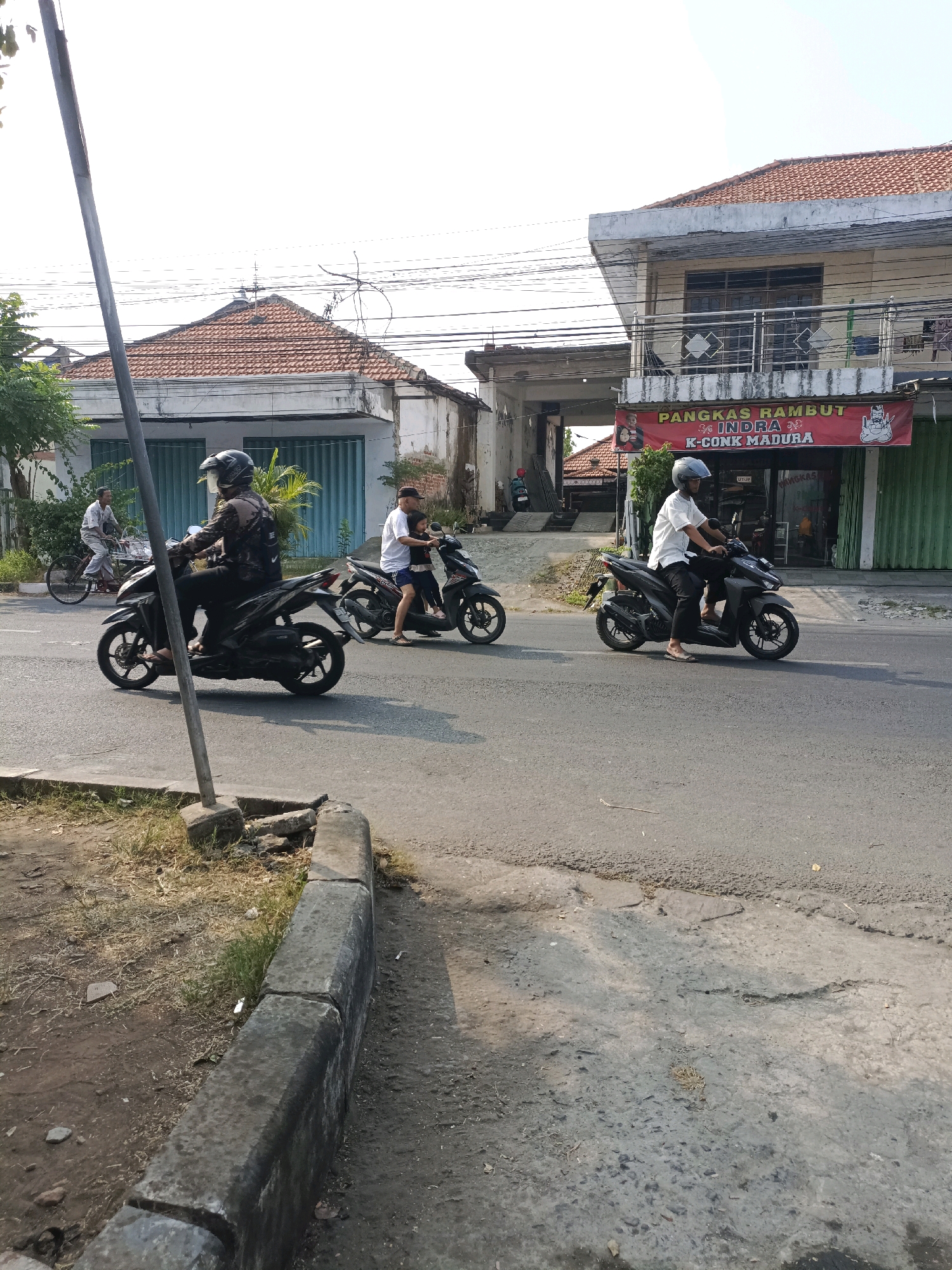 In Sukorejo, Indonesia, a street scene unfolds under a bright sky. Three motorcycles are on the road, with two riders clearly visible. The first motorcycle, a dark color, is ridden by a person wearing a patterned jacket and a helmet. To their right, a second dark motorcycle carries two individuals, a driver and a passenger, both wearing white shirts and helmets. Further down the road, a third dark motorcycle is partially visible. A man on a bicycle rides in the background on the left side of the image, heading away from the camera. Buildings line the street; one building features a red banner with text in Indonesian. The road surface appears paved with some wear and tear. The environment is characterized by a sunny day and a typical urban street setting in Indonesia.