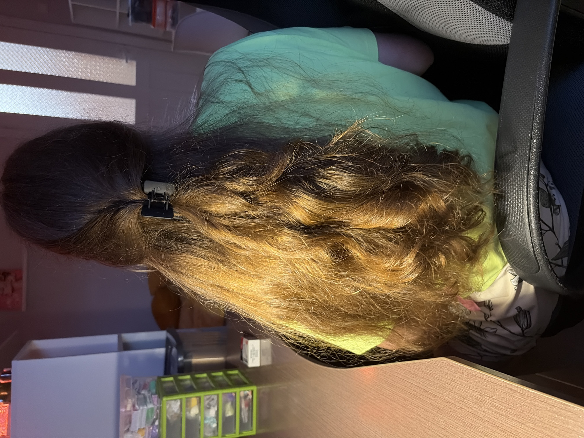 The image shows the back of a person's head and shoulders, with their long, wavy, brown and blonde hair prominently featured. The hair is held back with a black hair clip. The person is wearing a light green t-shirt. They are sitting in a black office chair with mesh detailing and a black armrest. The background is slightly blurred, but hints of a room interior are visible, including shelves and some boxes or containers on a desk. The lighting suggests it might be daytime, with some light casting shadows on the person's hair, possibly from blinds or a window. The overall scene appears to be an indoor, casual setting, possibly a home office or study area. There is no visible text in the image.