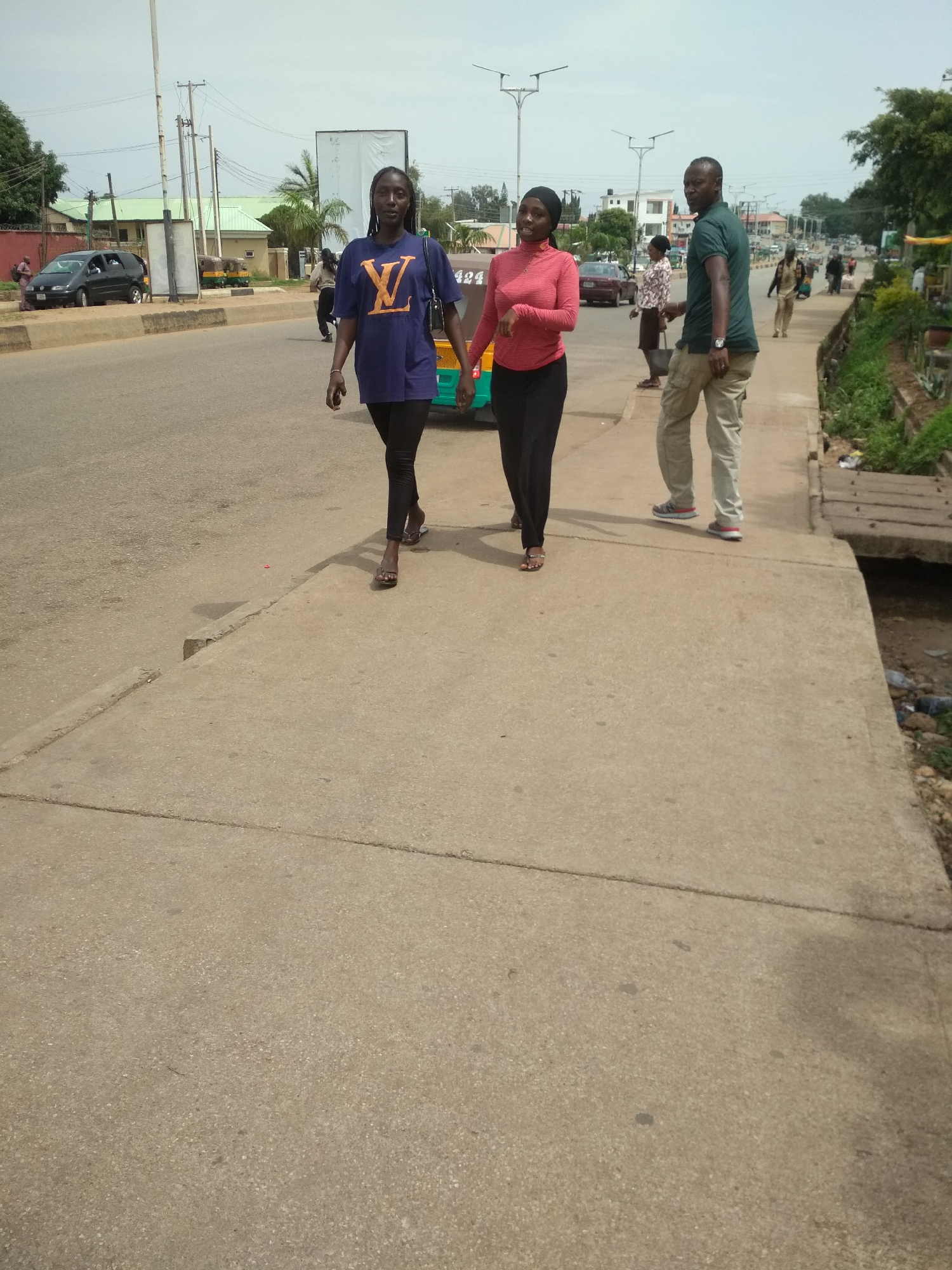 Two women walk side-by-side on a concrete sidewalk adjacent to a paved road in Jos, Nigeria. The woman on the left wears a dark blue t-shirt with a large yellow "LV" logo and black leggings. The woman on the right wears a coral pink long-sleeved shirt and black leggings. Both are walking towards the viewer's left. Behind them, a man in a dark green t-shirt and tan pants walks away from the viewer. In the background, there are several cars, motorcycles, and buildings, including a large white billboard. The scene appears to be a typical street in the city, with power lines and streetlights visible.