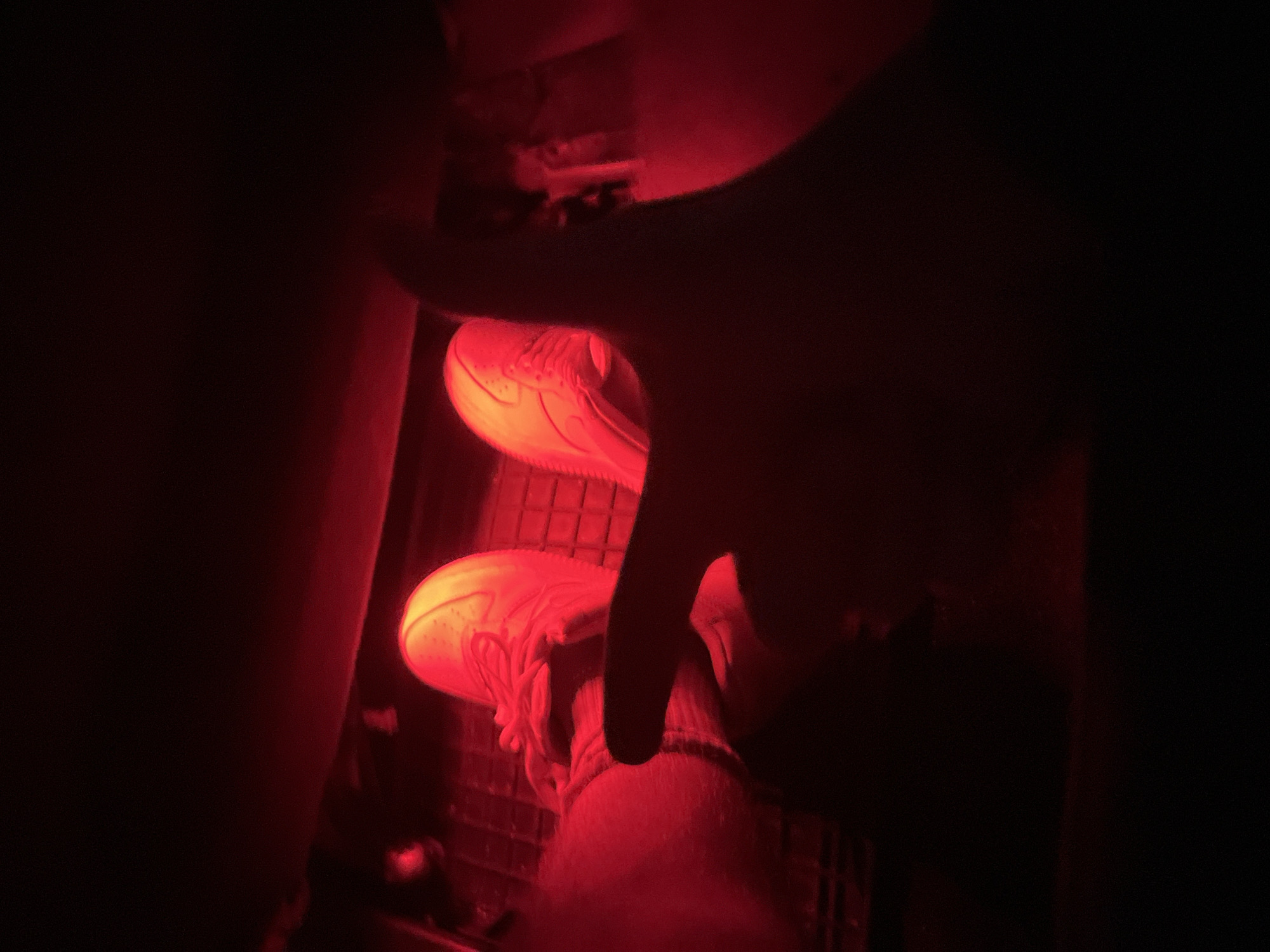 The image shows a close-up view of a person's feet wearing red-lit sneakers, with the right foot on a car pedal, likely the accelerator. A hand is visible in the foreground, partially obscuring the view of the feet and pedals. The lighting is predominantly red, suggesting an interior car environment with red ambient lighting. The location in the city of Zlatograd, Bulgaria, is not discernable from the image itself.