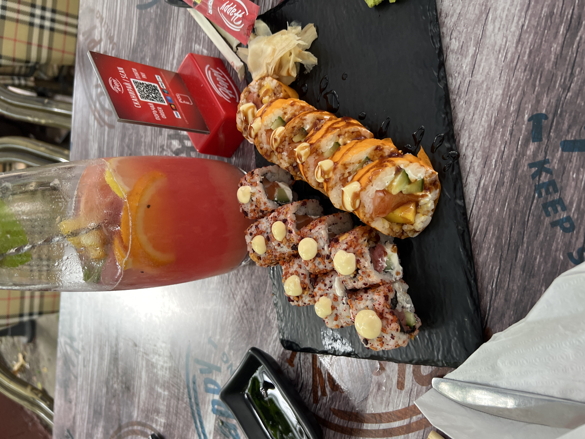 A table in Burgas, Bulgaria, is set with a meal. The table is a textured wood-look, with a partial "Keep" showing on the edge, and is set outdoors. A glass of red-tinted beverage with citrus slices is on the left of the frame next to a red table marker. Adjacent to the beverage sits a black tray of sushi rolls with ginger and avocado on the right edge. Some of the sushi rolls are topped with cream-colored sauce, and a small black container holds a dark liquid on the lower left edge. There is also a napkin and silverware on the right edge of the image.