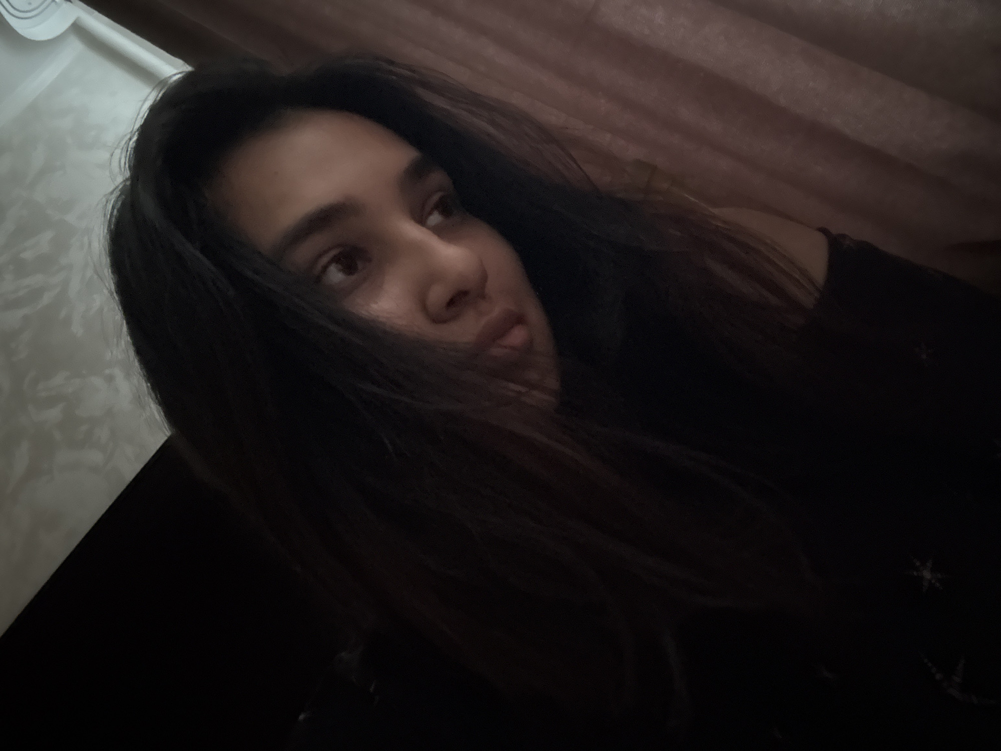 A young woman with dark hair is shown in a dimly lit indoor setting, possibly a bedroom, with a curtain in the background. The lighting is low, creating shadows and a soft focus on her features. Her gaze is directed away from the camera, with her head tilted to the side. The image is a close-up, focusing on her face and upper body. The background is indistinct due to the low light conditions. The provided location, Misaylovo, Russia, is not discernible from the image itself.